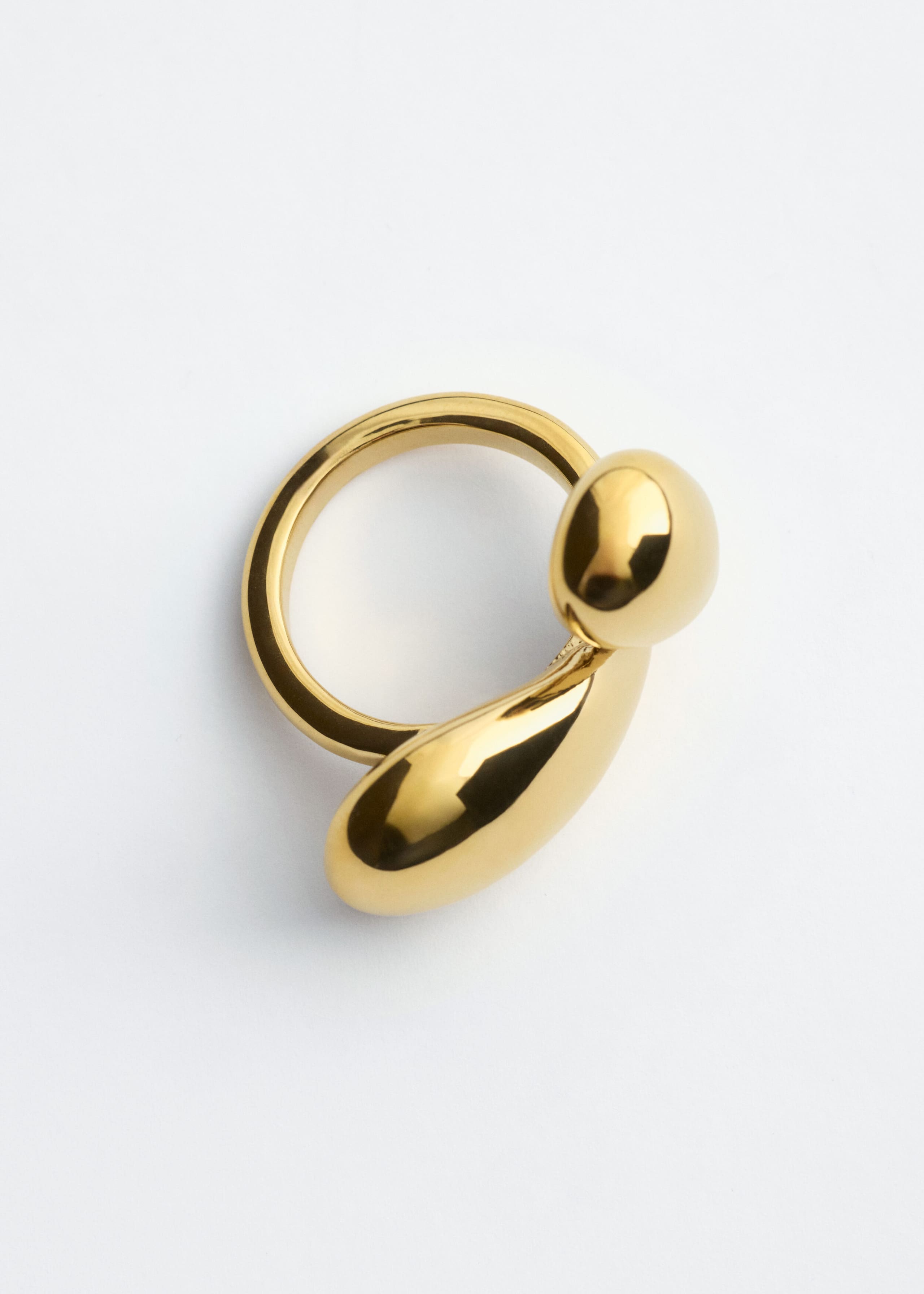 Un anello in ottone color oro presenta una finitura altamente lucida e riflettente e un design scultoreo con due forme fluide e bulbose. Visto frontalmente, il giro anello si curva in una piccola goccia arrotondata e in una forma a goccia allungata più גדולה.
