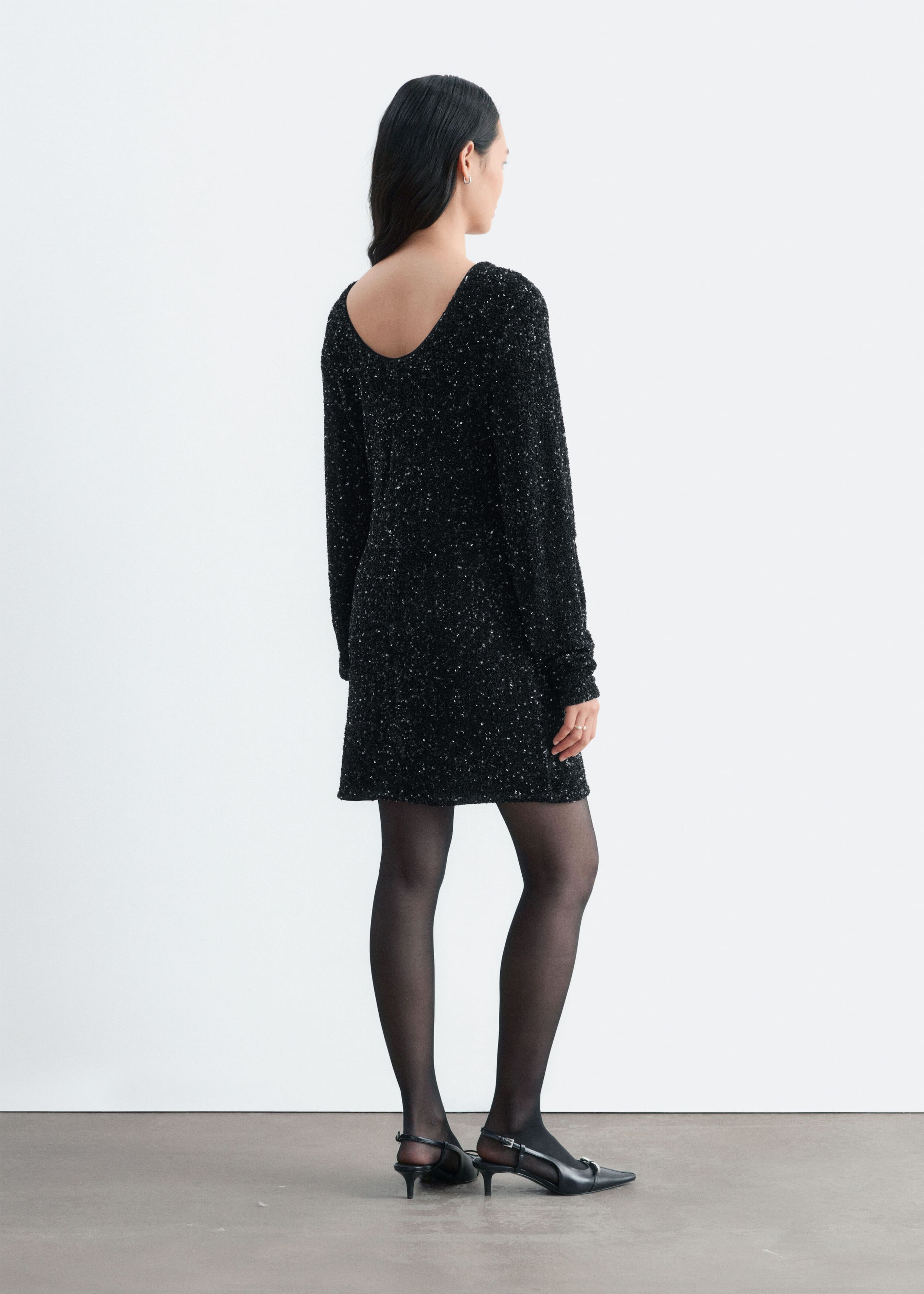 The model is wearing a short black sequined mesh dress with long sleeves and a scooped back neckline. The dress is styled with sheer black tights and black pointed-toe slingback shoes.