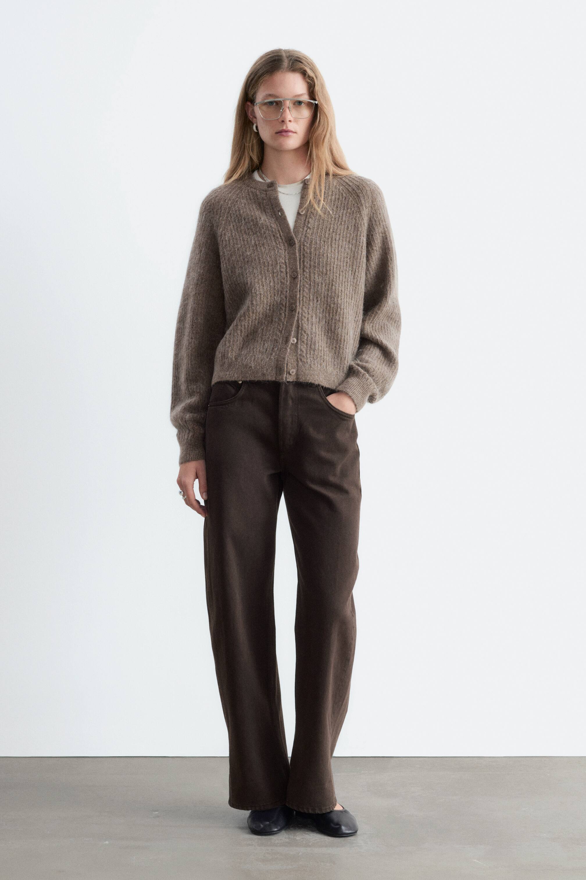 Agrandir l'image: Un cardigan en mélange de mohair de couleur taupe brun est présenté de face, avec une coupe décontractée, des manches raglan et une patte de boutonnage ornée de détails en tricot torsadé. Le cardigan est porté sur un t-shirt blanc et associé à un pantalon large brun foncé, mettant en valeur son col, ses poignets et son ourlet côtelés.
