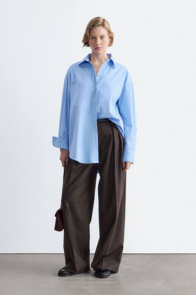 Wool-blend tailored trousers in dark brown are being worn by the model, featuring a wide-leg silhouette, front pleats, and pressed creases. The trousers are styled with a thin black belt and a partially tucked, oversized light blue button-up shirt. - 1