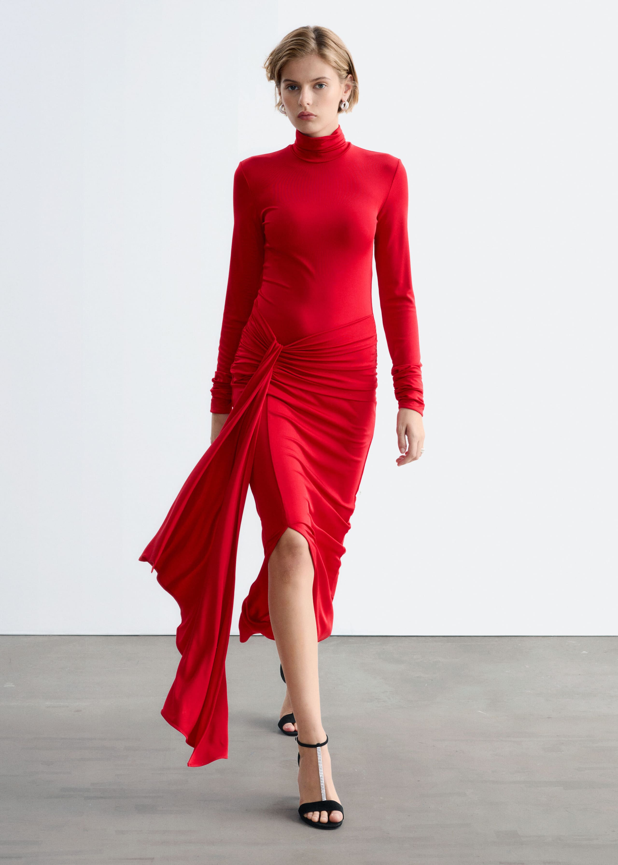 A red jersey dress featuring a mock neck and long sleeves is being worn by the model walking forwards. The slim-fit dress is styled with black high-heeled sandals and features a draped tie detail at the waist with a long, flowing panel and a front leg slit.