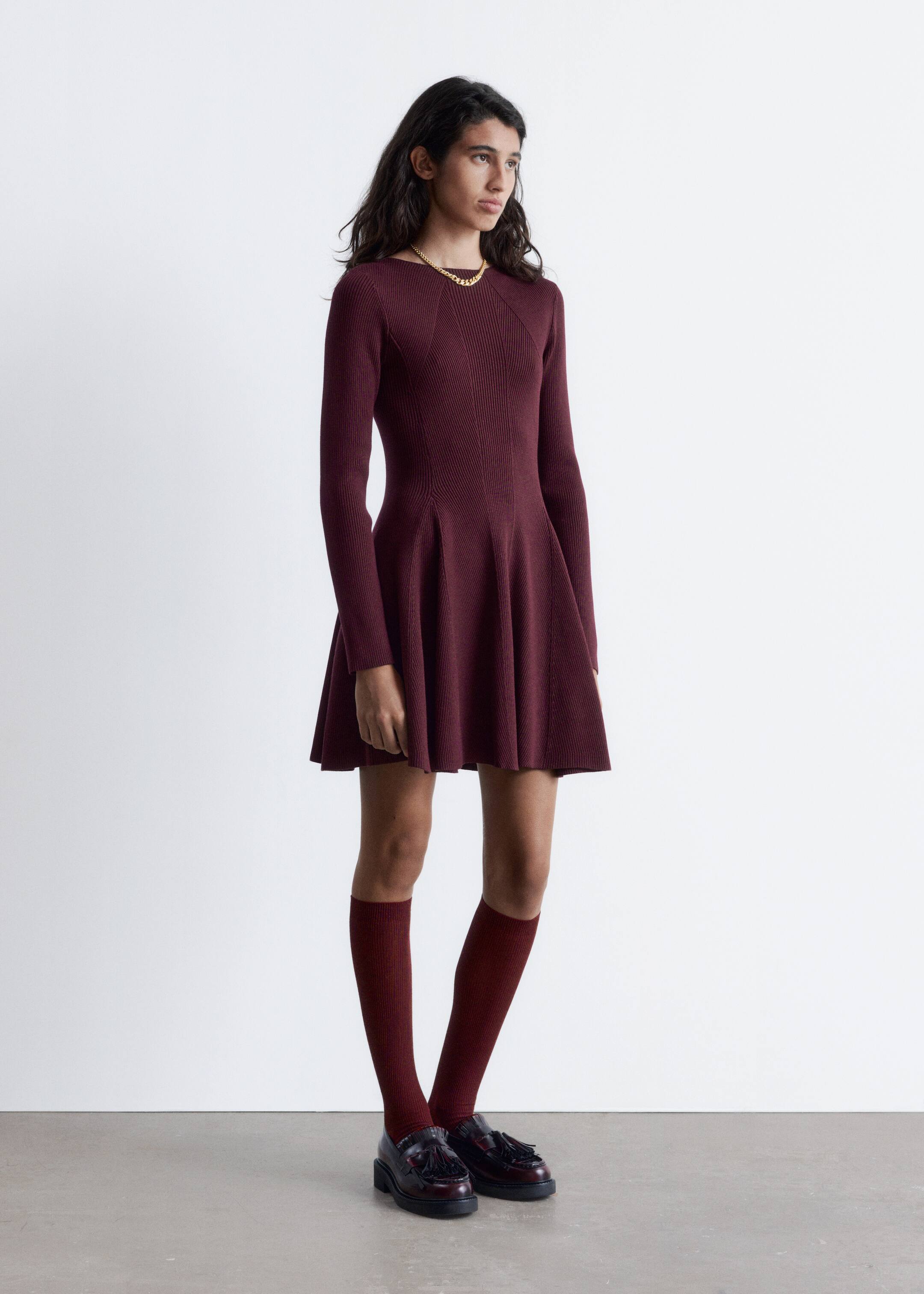 Agrandir l'image: Une robe mini en tricot bordeaux avec une finition côtelée, dotée d'un col large et haut, de manches longues, d'une taille ajustée et d'une jupe évasée à panneaux, est portée par le mannequin. Le mannequin porte également un collier chaîne doré, des chaussettes montantes bordeaux assorties et des mocassins foncés.