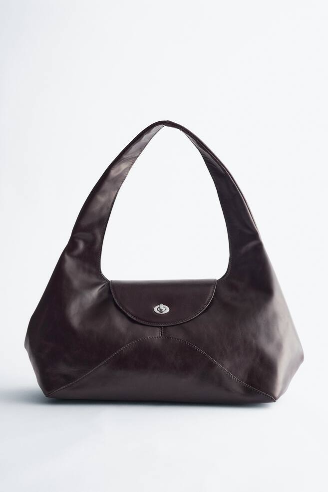 A dark brown suede shoulder bag is facing front, featuring a curved, sculptural silhouette and a wide, sloped shoulder strap. It is displaying a front flap with a silver-toned twist-lock fastening and decorative curved cut-line seams. - 1