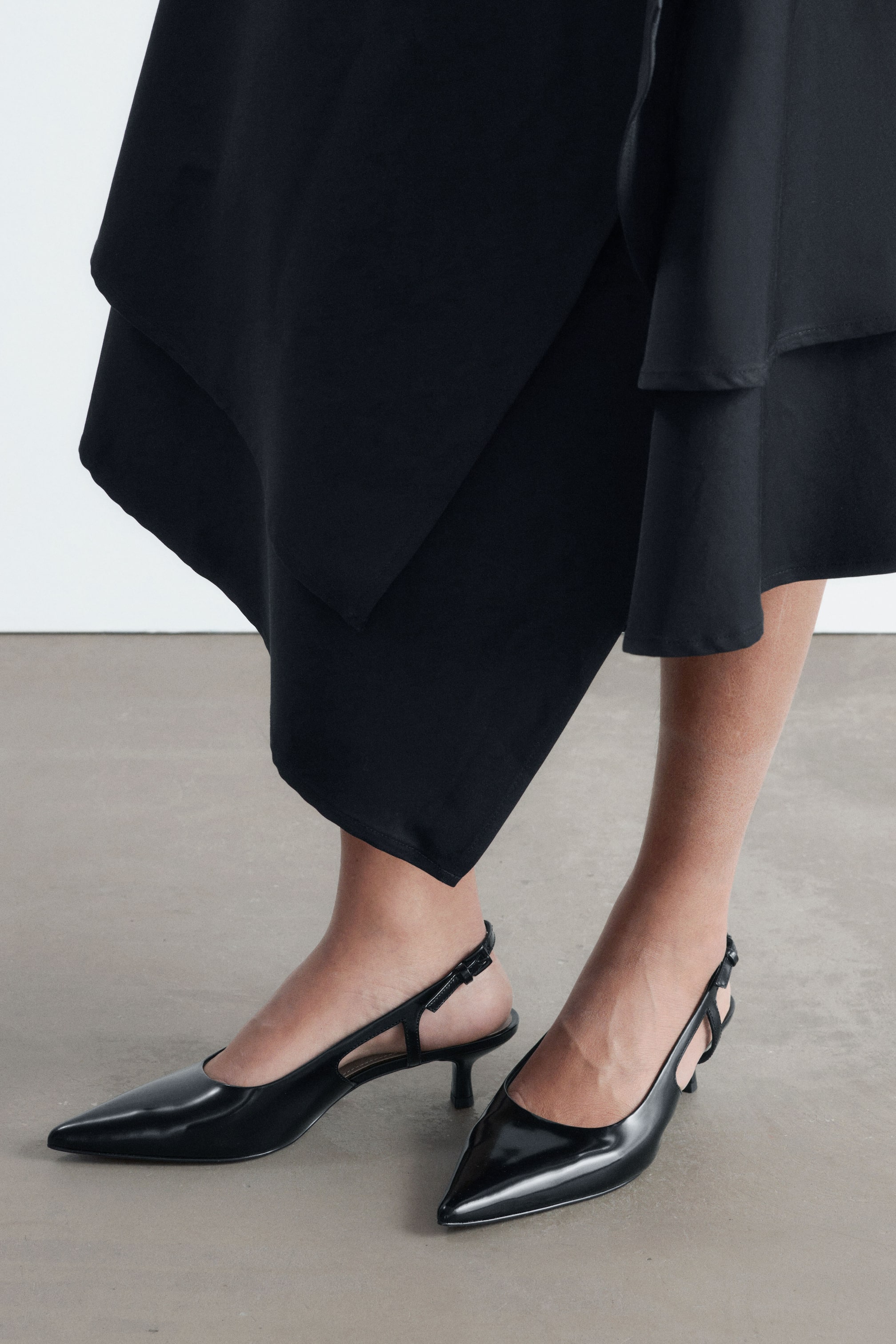 Grösseres Bild ansehen: Schwarze Slingback-Schuhe aus Lackleder mit spitzer Spitze, Kitten Heels und schmalen Fersenriemen mit verstellbarer Schnalle werden vom Model getragen.