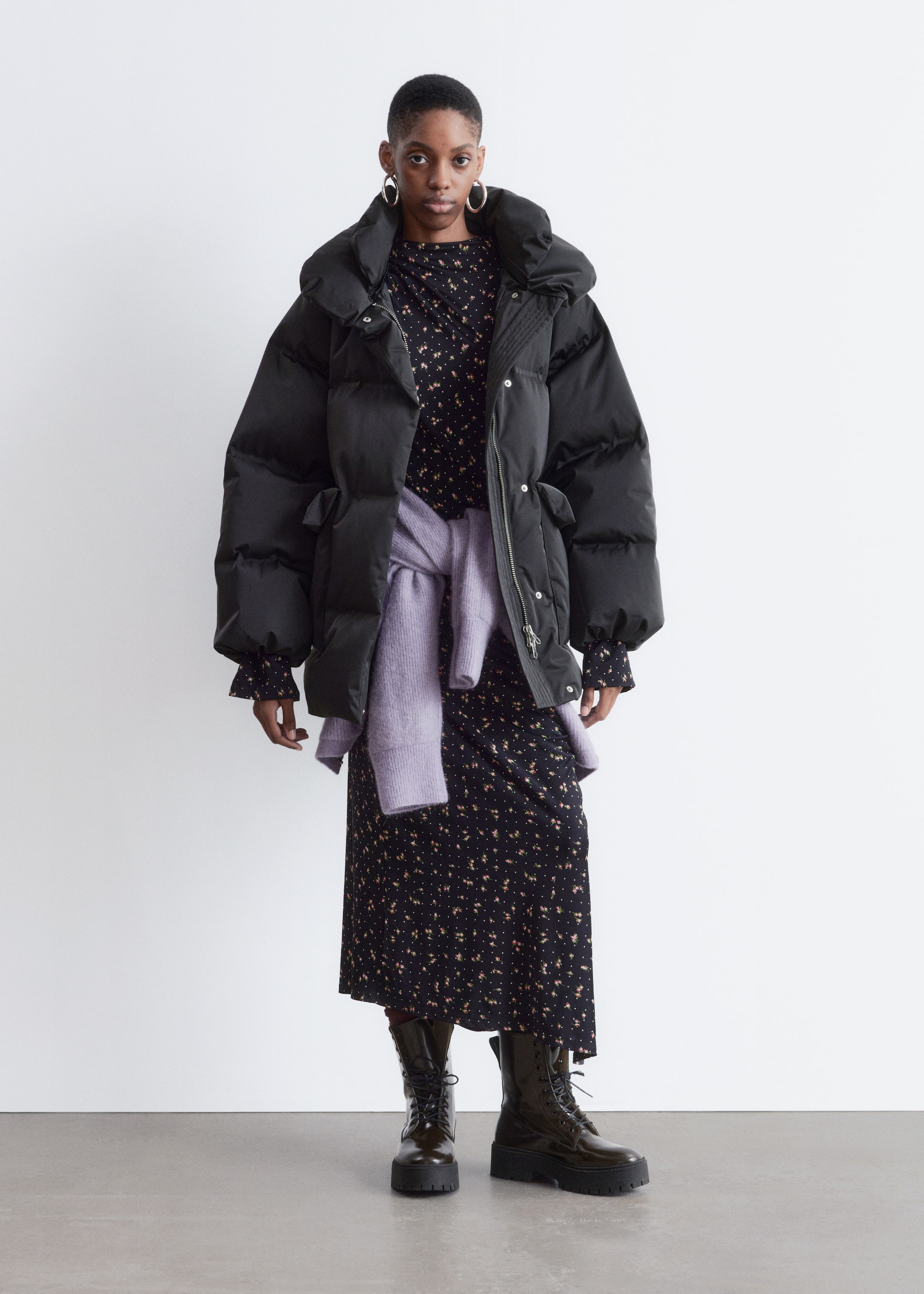 View larger image: A black puffer coat is being worn, facing forward. It is designed with an oversized, voluminous silhouette, featuring a high stand collar, a front zipper closure, and two ruched flap pockets. The coat's long sleeves are revealing the patterned cuffs of a garment worn underneath.