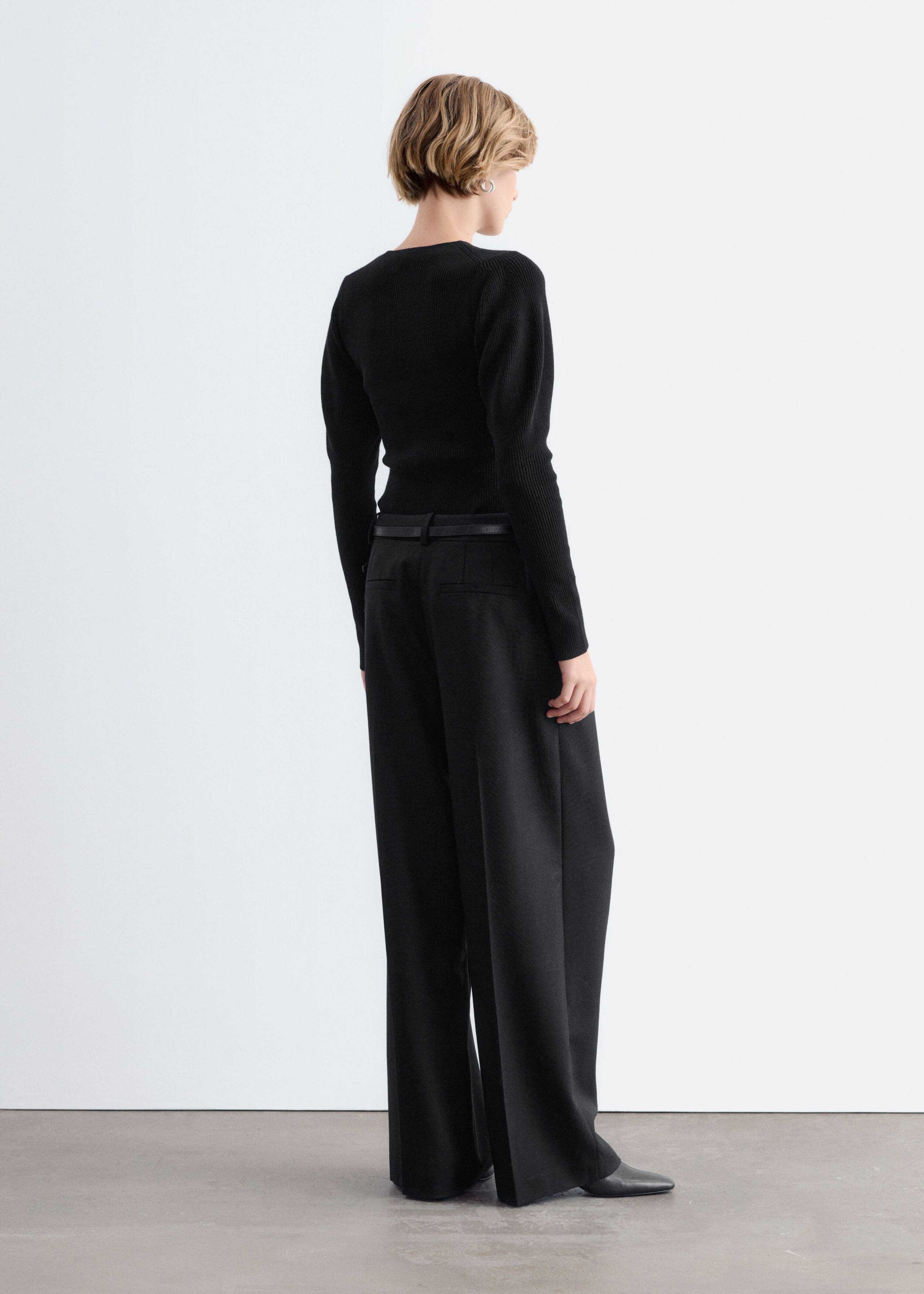 A black rib-knit top with long sleeves and softly puffed shoulders is worn by the model, who is facing away from the camera. The fitted top is styled tucked into black wide-leg trousers with a thin black belt.
