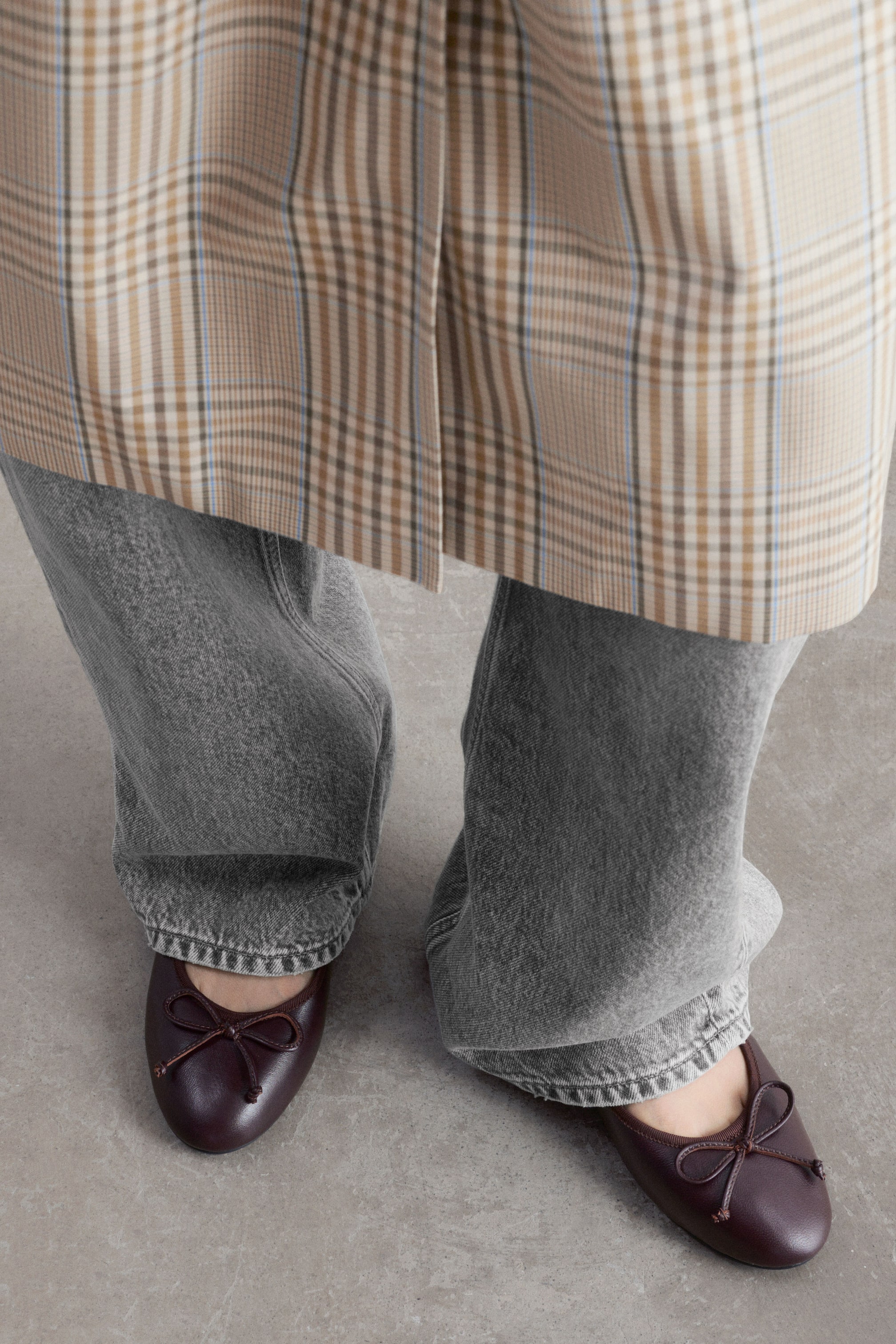 Agrandir l'image: Des ballerines en cuir d’un bordeaux profond, avec des bouts arrondis agrémentés de petits nœuds, sont portées par le mannequin. Les ballerines sont associées à un jean en denim gris à jambe large et à un long manteau à motif à carreaux.