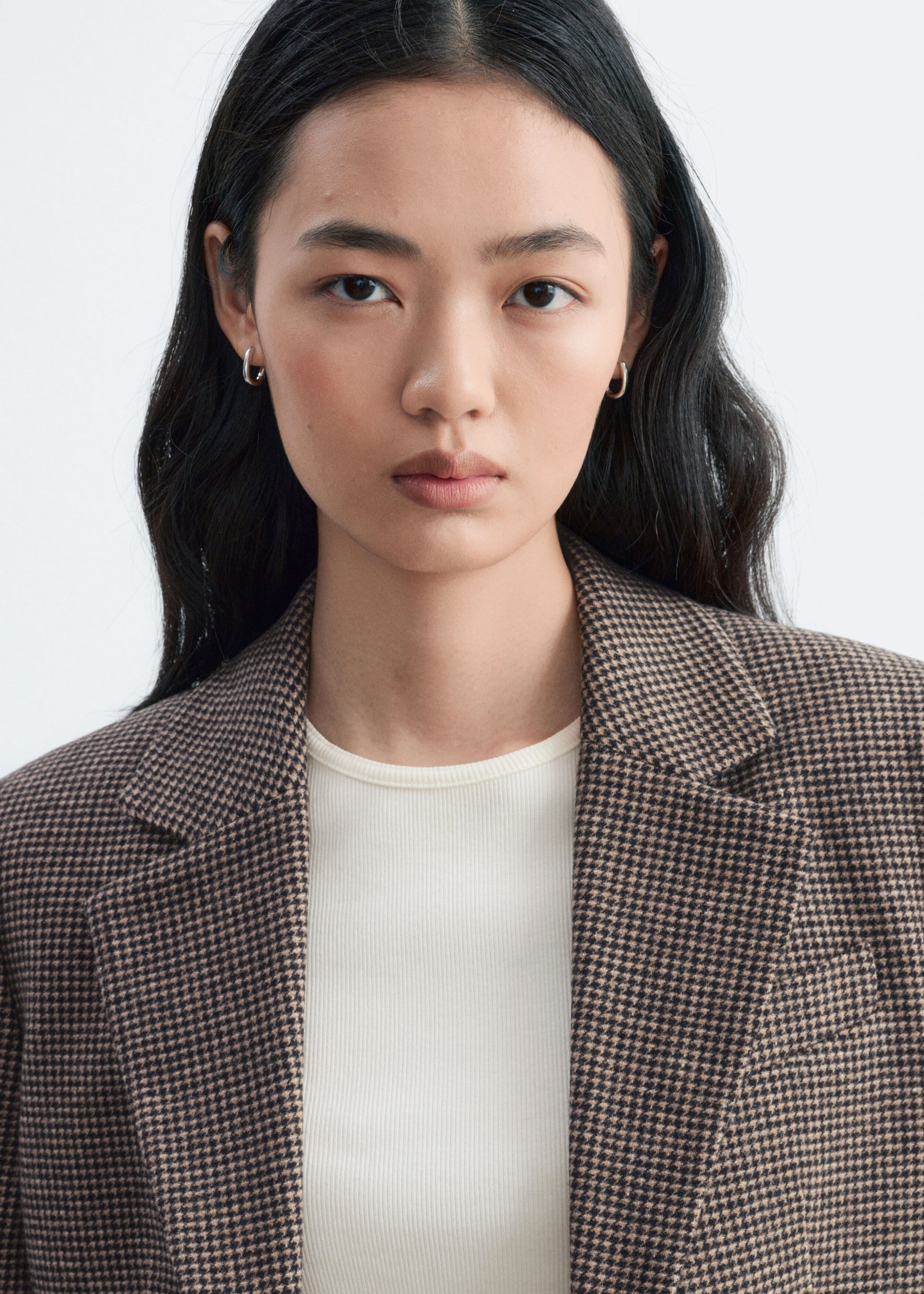 A houndstooth blazer in a brown and black pattern is being worn by the model over a white ribbed top, showcasing its notch lapels and textured wool-blend fabric.
