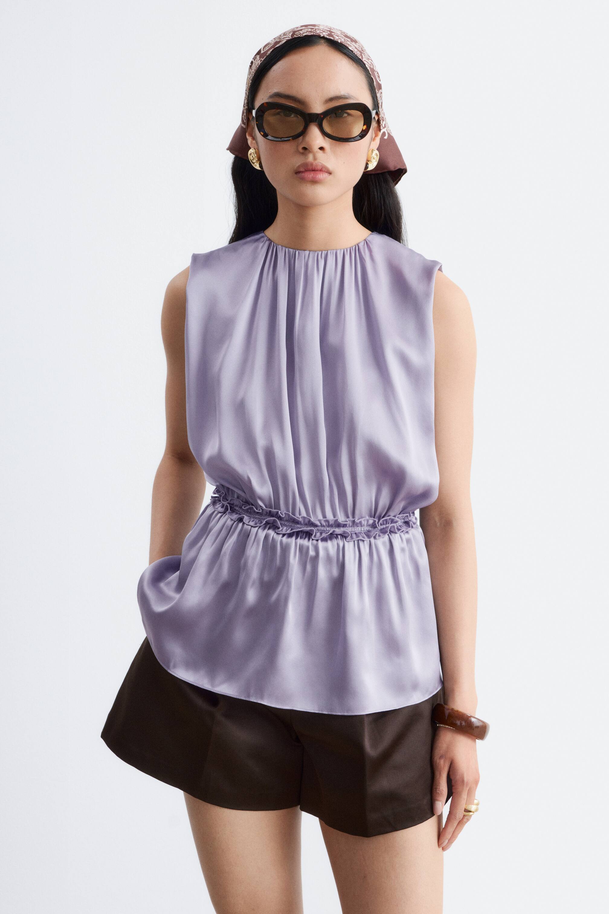 Agrandir l'image: Un haut sans manches en satin lilas présente un col montant froncé et une taille élastiquée, créant une silhouette subtile en péplum.
