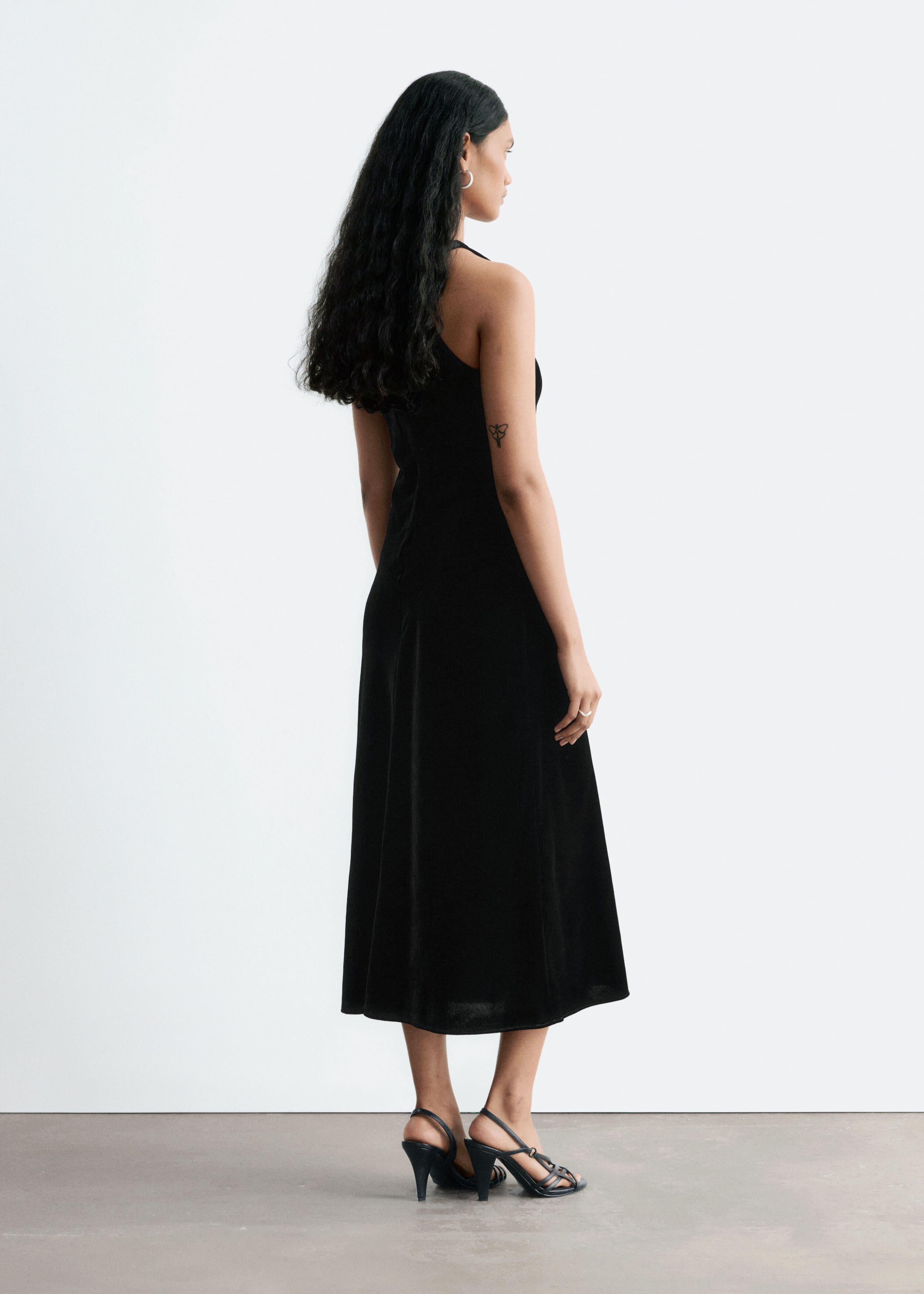 A black sleeveless midi dress with a subtle satin finish is worn by the model, shown from a three-quarter rear angle. The dress features a round neckline and an A-line silhouette, styled with black strappy high-heeled sandals.