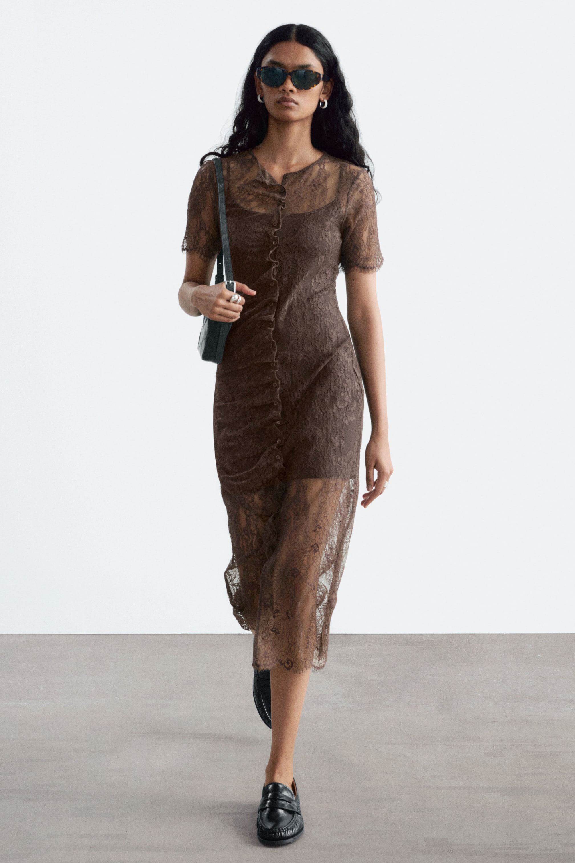 Größeres Bild ansehen: Ein braunes Spitzenkleid mit schlanker Silhouette wird von dem Model getragen, das im Gehen eine schwarze Schultertasche trägt. Das Kleid verfügt über kurze Ärmel mit gewellten Bündchen, eine vorderseitige Knopfleiste mit Raffung sowie einen Midi-Rock mit gewelltem Saum.