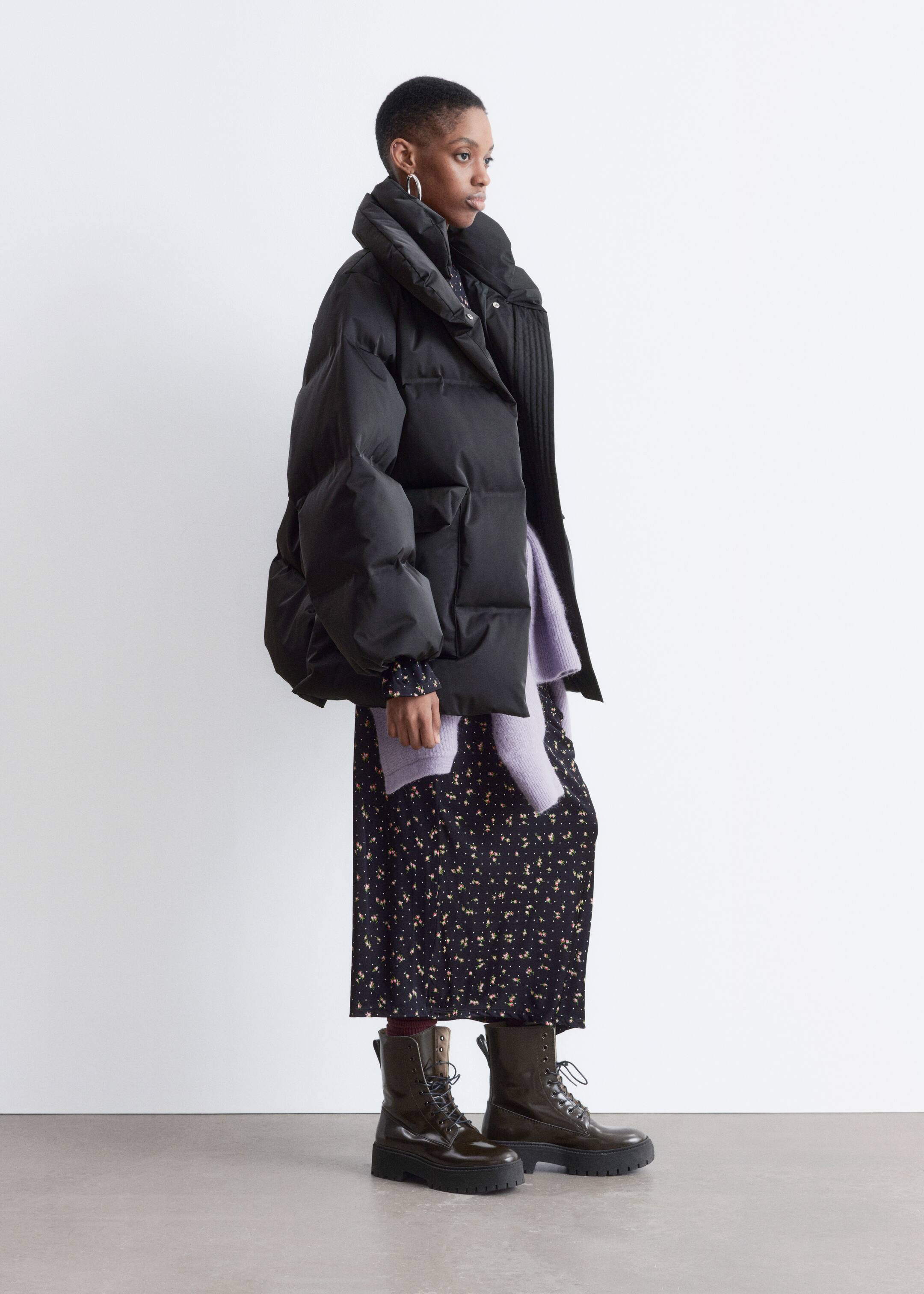 View larger image: A black puffer jacket is being shown from a side angle, featuring a voluminous design with a high, wide collar and visible snap buttons on its front. A ribbed panel is visible at the inner collar area, and a light purple sweater is layering underneath the jacket.