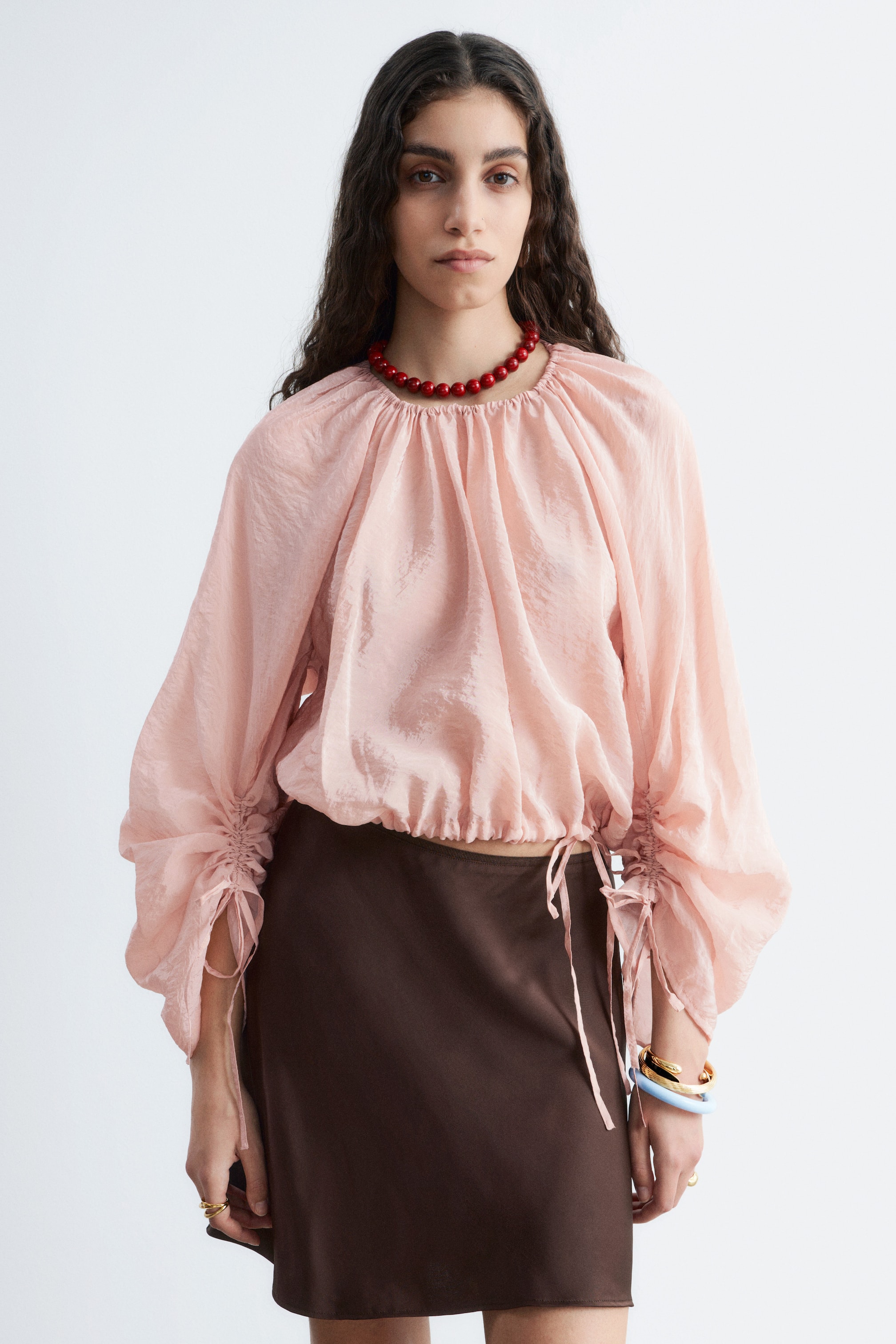 Visa större bild: En ljusrosa blus med skrynklig textur, en rynkad rund hals och voluminösa långa ärmar med rynkade dragsko-detaljer. Blusen har även en elastisk fåll och är stylad med en mörkbrun satinliknande minikjol.