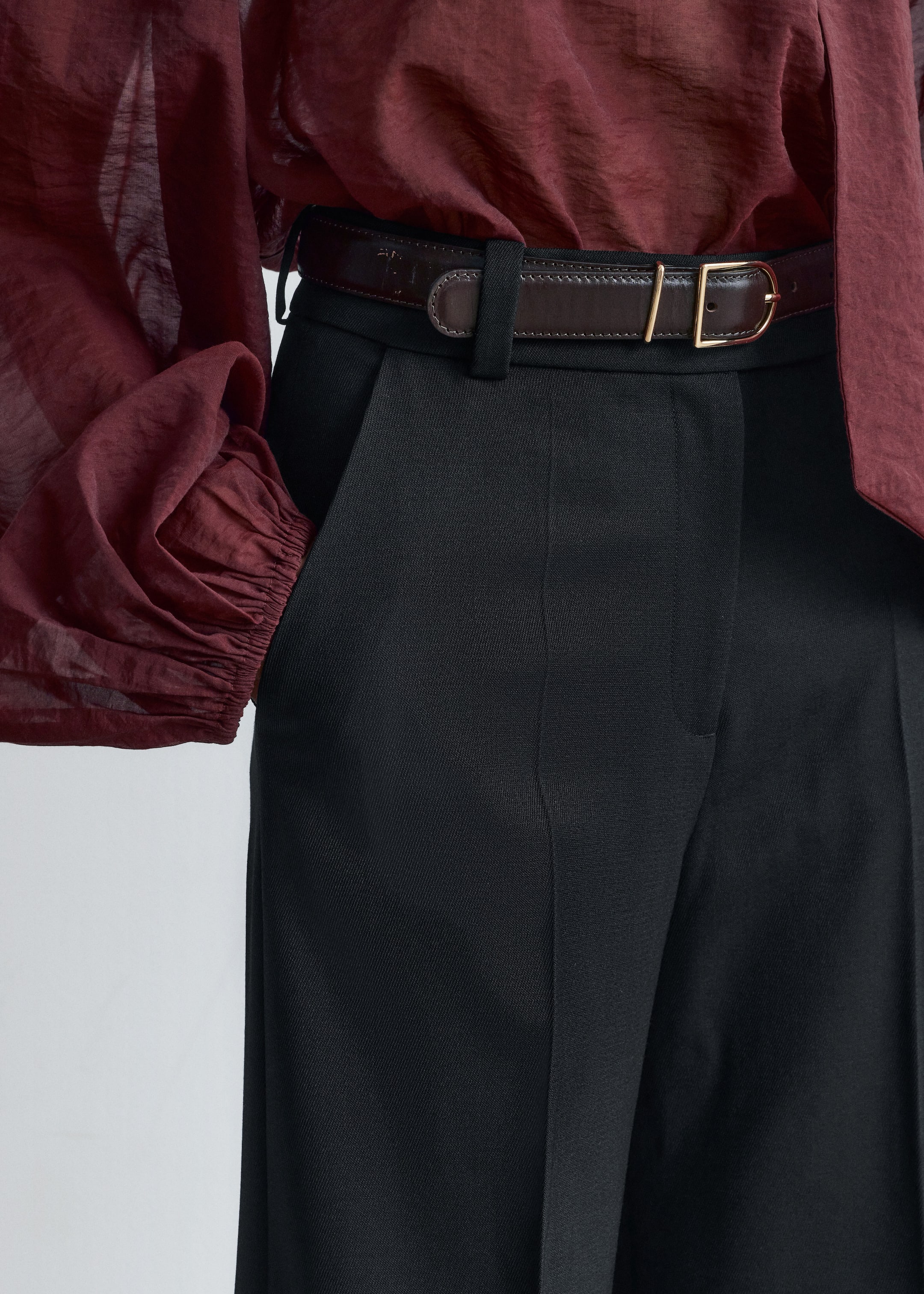 Visualizza immagine più grande: Pantaloni neri, che mostrano una sottile trama a spina di pesce, presentano una piega frontale prominente e passanti per cintura. Una cintura in pelle marrone scuro con una fibbia rettangolare color oro stringe la vita, sopra la quale è drappeggiata una camicetta trasparente bordeaux con maniche voluminose e arricciate.