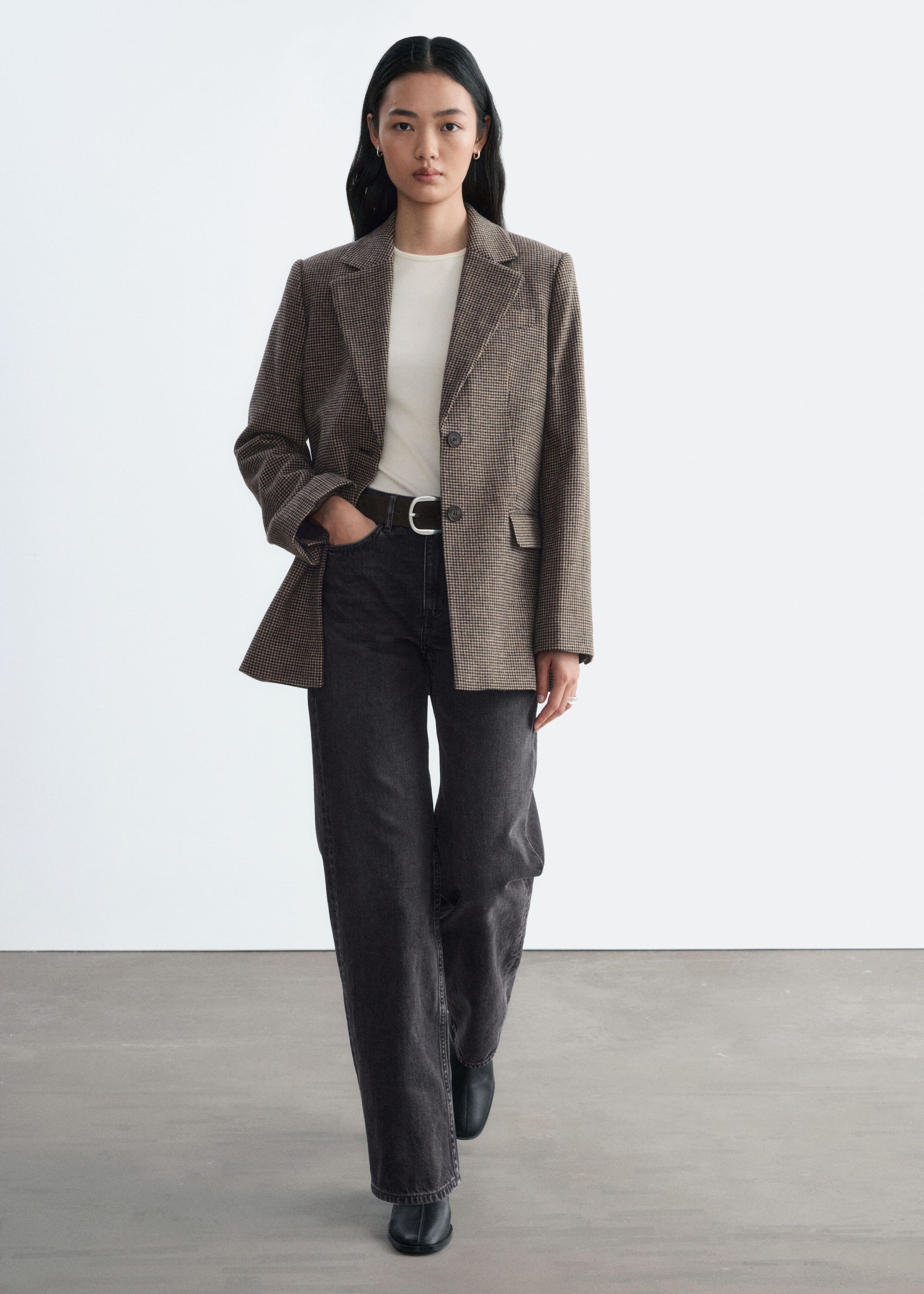 A brown houndstooth-patterned single-breasted blazer is worn open by the model over a cream-coloured crew-neck top and dark grey wide-leg jeans. The blazer features notched lapels, front buttons, and flap pockets.
