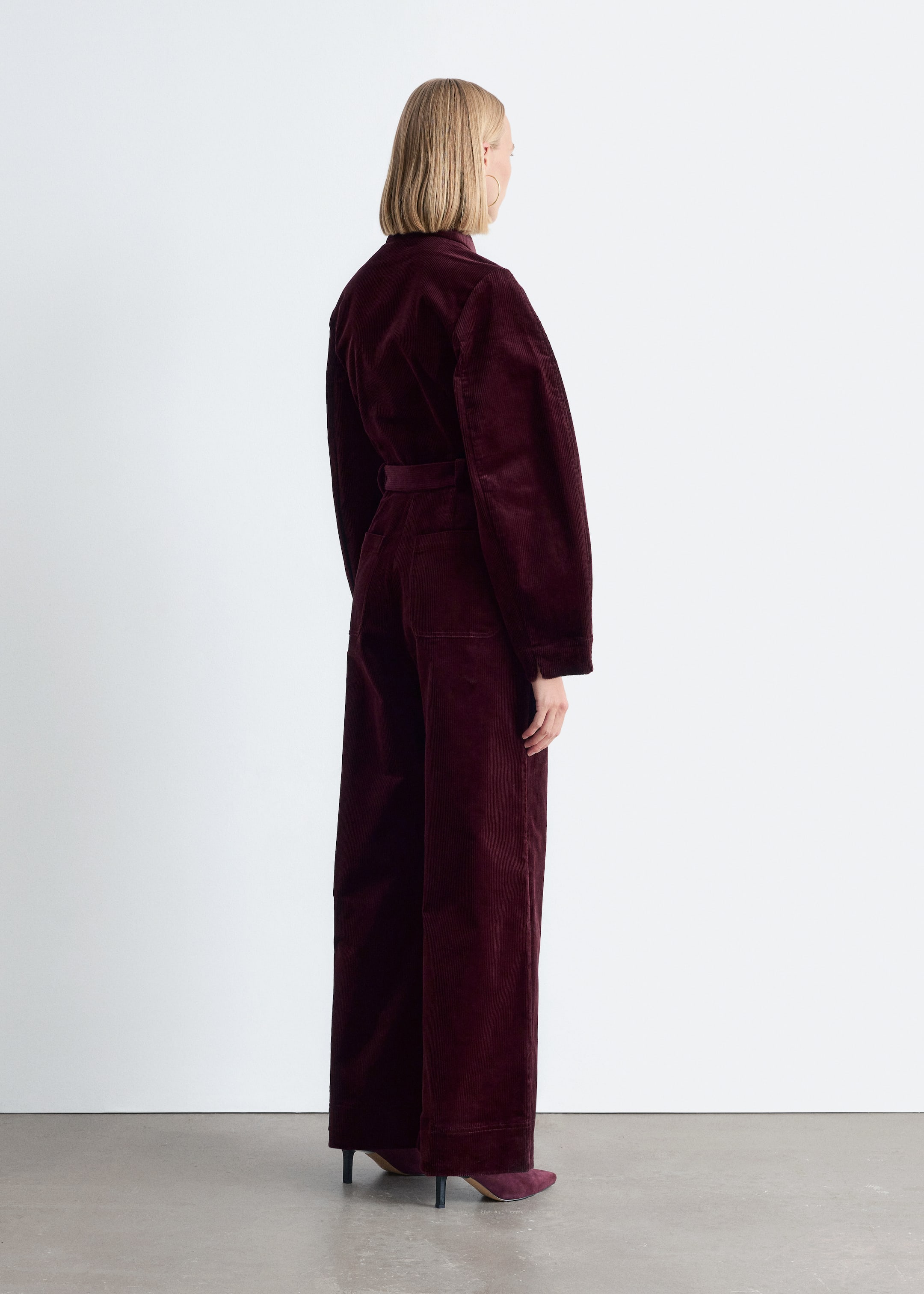 Größeres Bild anzeigen: Ein dunkelroter, hoch taillierter Baumwoll-Cord-Jumpsuit wird vom Model getragen, mit langen Ärmeln und einem Bindegürtel, der die Taille am Rücken betont. Passende burgunderrote Absätze werden ebenfalls getragen.