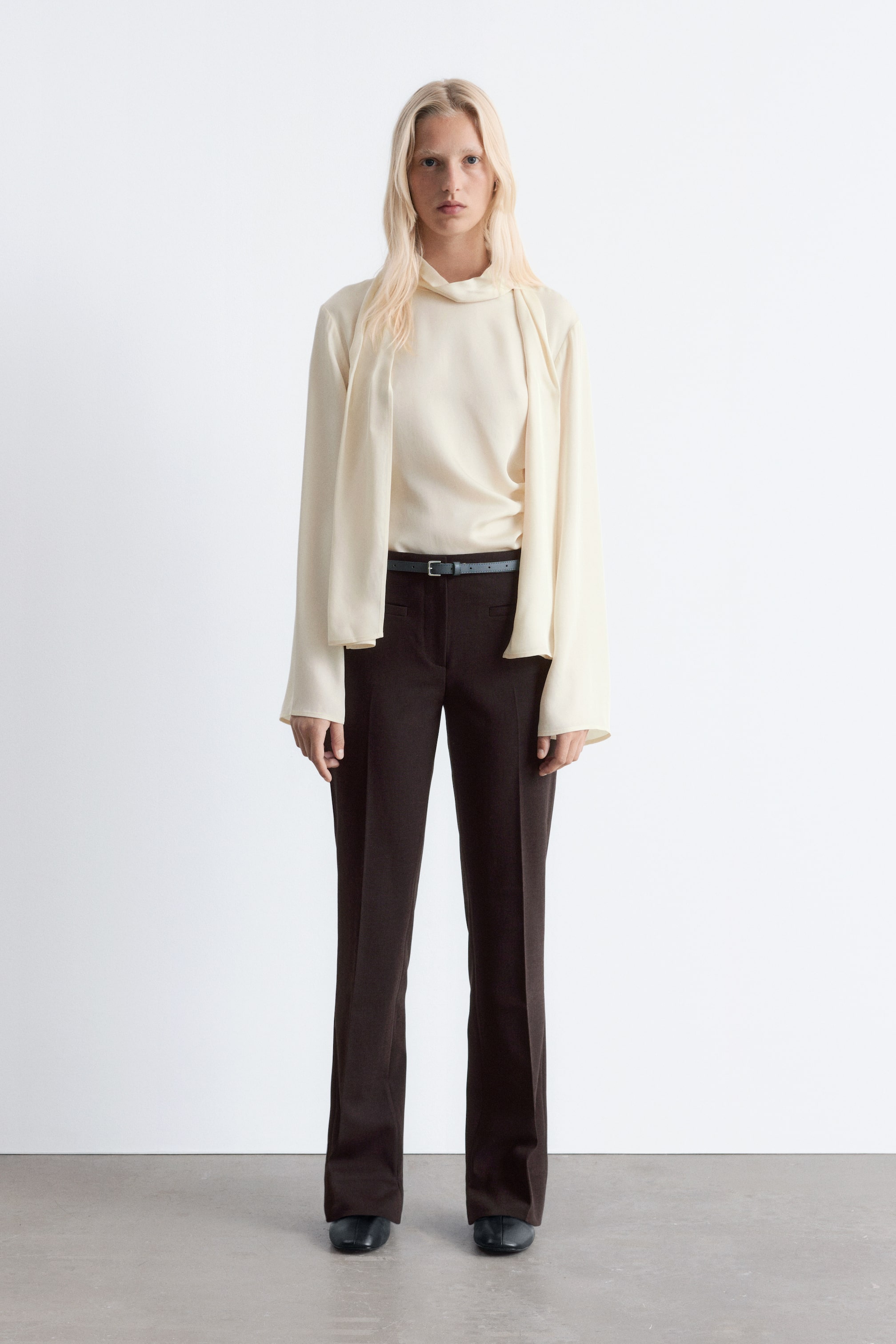 Ver imagem maior: Um top de manga comprida creme com um decote alto drapeado e mangas largas e fluídas está a ser usado com calças castanhas escuras de perna reta. Um cinto fino e escuro está a apertar as calças na cintura, que também apresentam um vinco frontal visível e estão a ser combinadas com sapatos escuros de bico fechado.