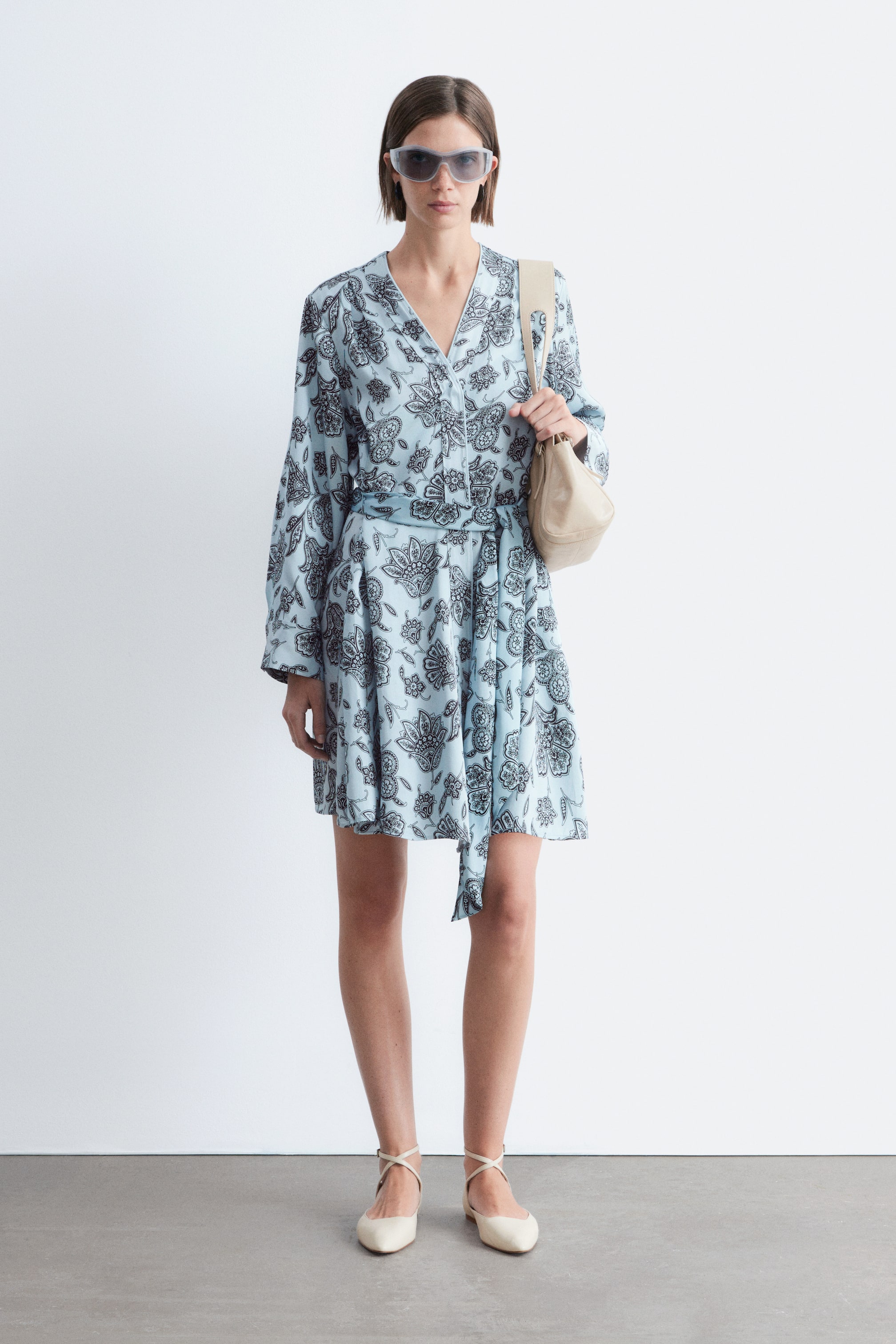 Grösseres Bild ansehen: Ein hellblaues Minikleid mit einem dunklen Paisley-Muster wird von vorne gezeigt, mit einem V-Ausschnitt und langen Ärmeln. Ein passender Bindegürtel betont die Taille, und der Rock fällt in einen kurzen Saum.