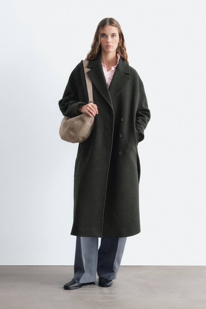 La modelo lleva un abrigo largo de doble botonadura en verde oscuro, con solapas de muesca y detalles de botones, sobre una camisa rosa claro y unos pantalones grises. La modelo lleva un bolso de hombro en topo y apoya una mano en el bolsillo lateral del abrigo. - 1