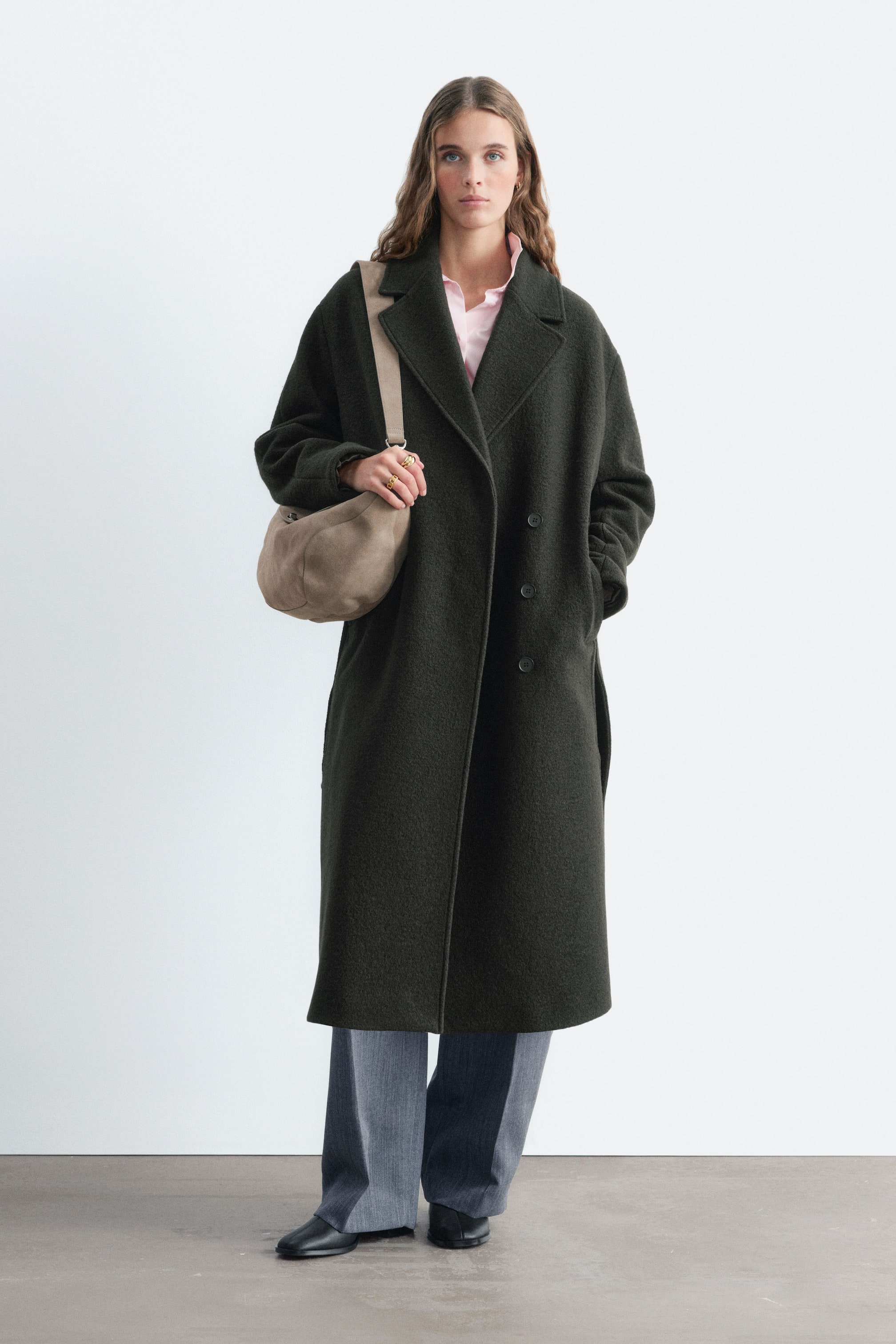 Ver imagen más grande: La modelo lleva un abrigo largo de doble botonadura en verde oscuro, con solapas de muesca y detalles de botones, sobre una camisa rosa claro y unos pantalones grises. La modelo lleva un bolso de hombro en topo y apoya una mano en el bolsillo lateral del abrigo.