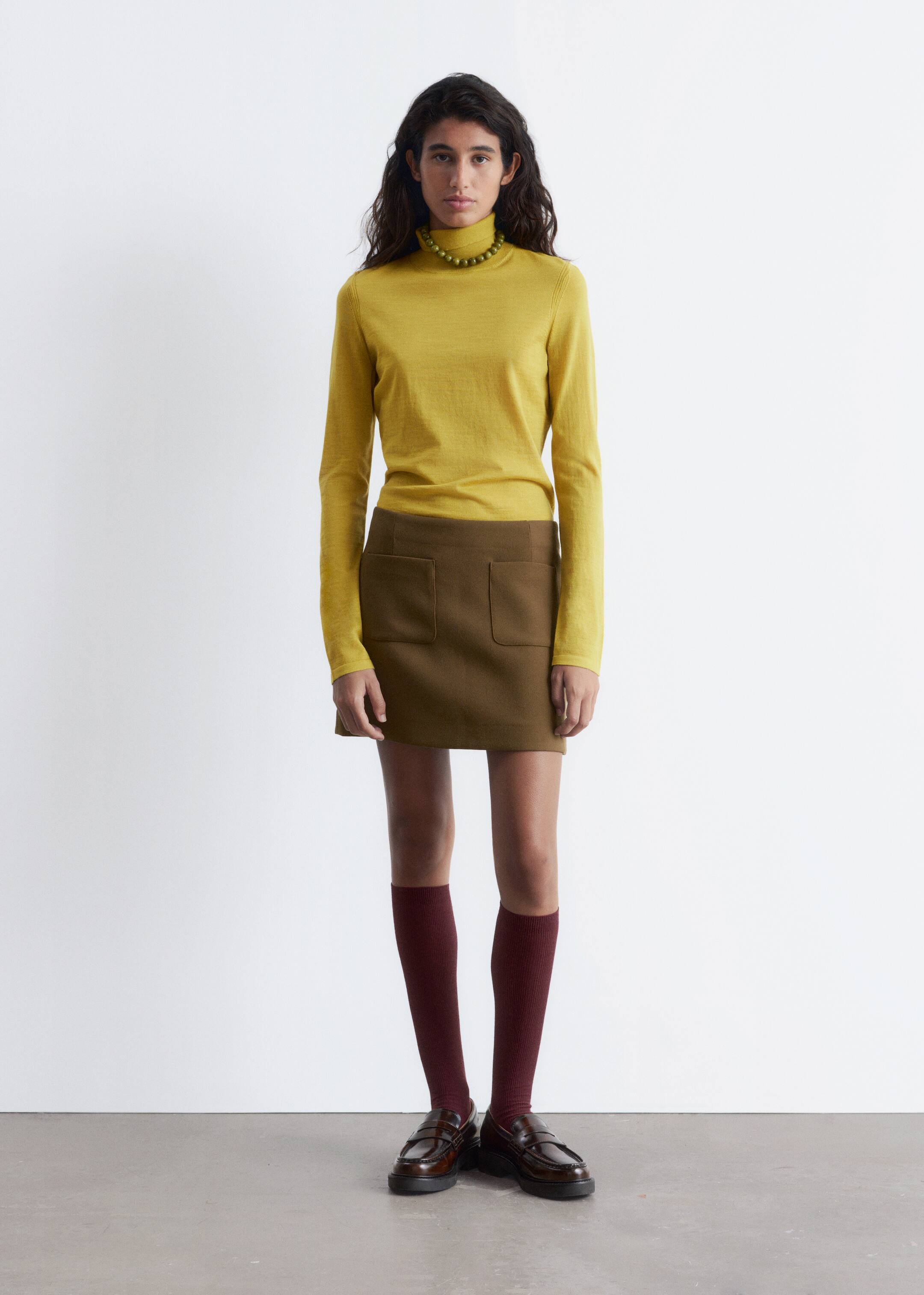 Ver imagen más grande: Una persona lleva un top de cuello alto de manga larga color chartreuse y una minifalda verde oliva, ambos presentados desde una vista frontal. La falda presenta dos grandes bolsillos de parche, y el conjunto se completa con un collar de cuentas verdes, calcetines burdeos hasta la rodilla y mocasines marrón oscuro.