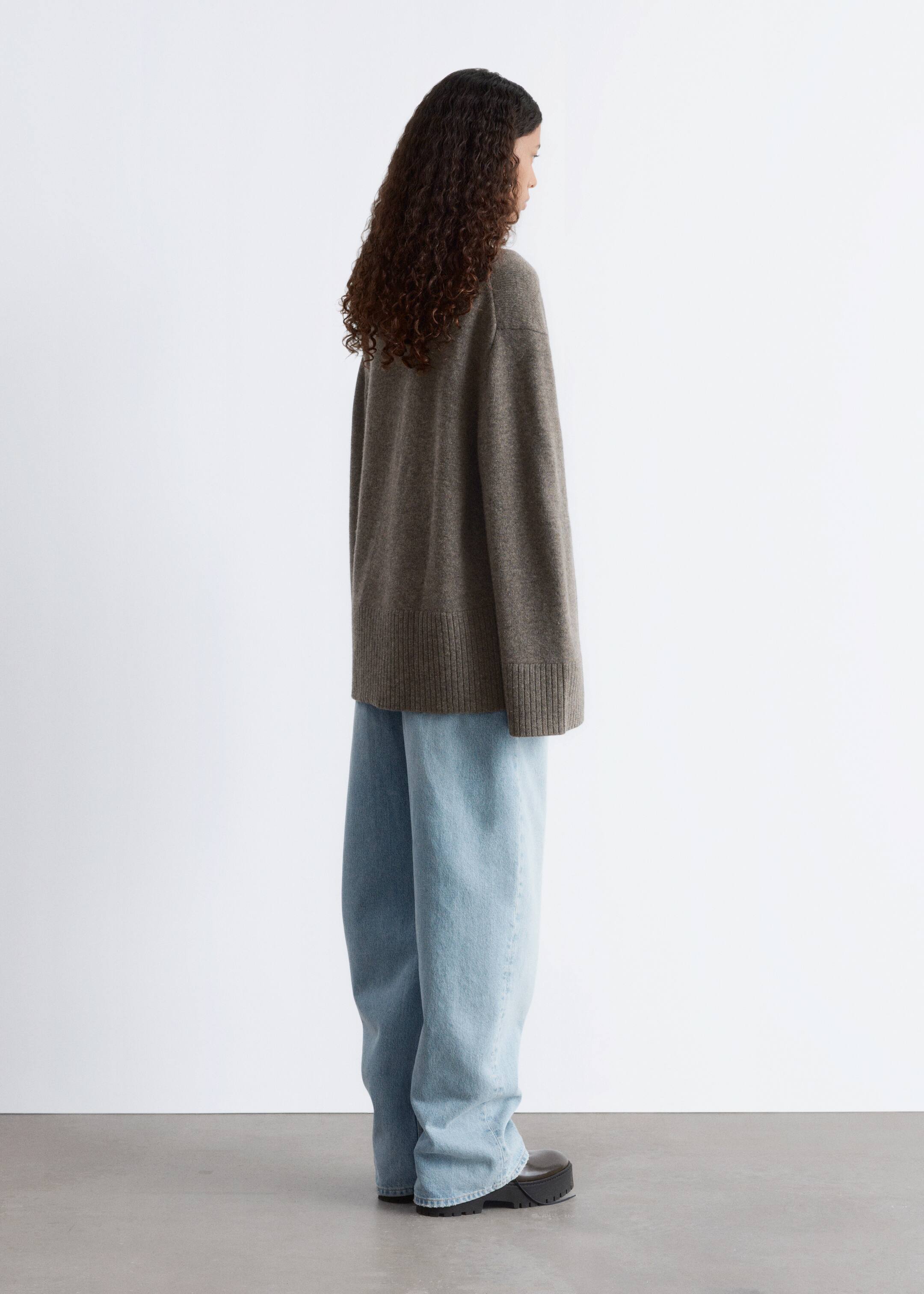 Grösseres Bild ansehen: Ein dunkelbrauner übergroßer Strickpullover wird in seitlicher Ansicht präsentiert, mit einer entspannten Silhouette und langen Ärmeln sowie gerippten Details am Saum und an den Bündchen, der über hellblauen Jeans im weiten Bein geschnitten getragen wird.