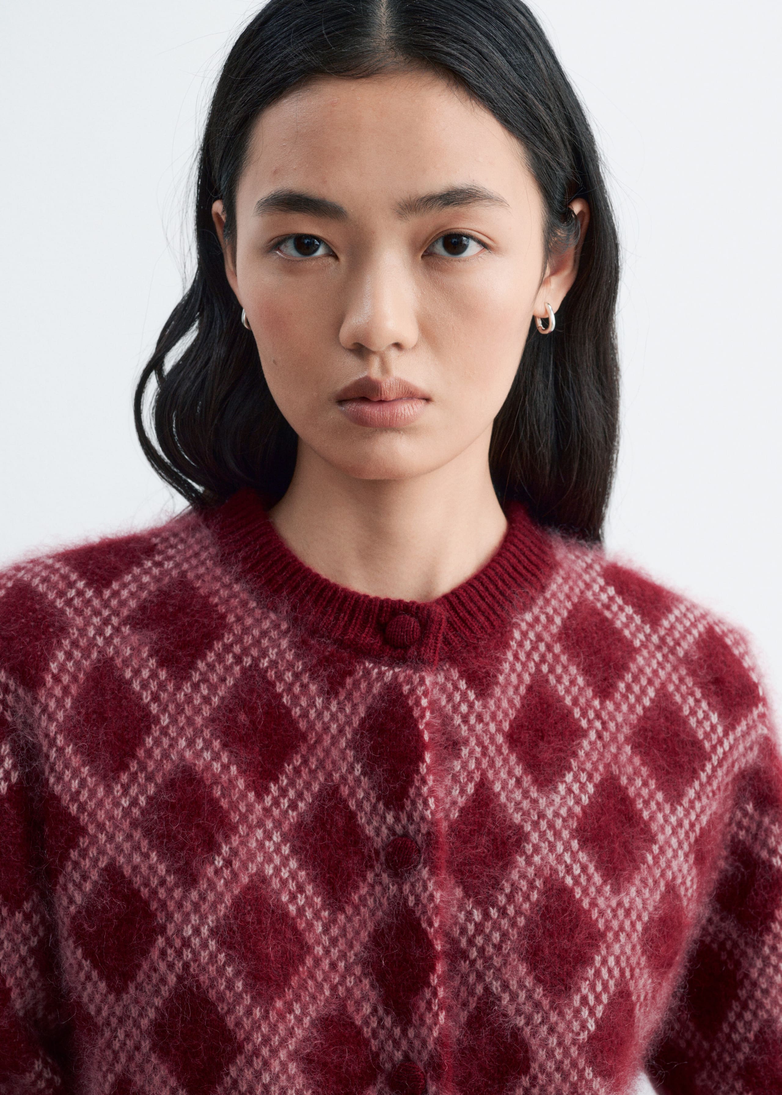 The model is wearing a fuzzy knit cardigan featuring a red and pink diamond pattern. The cardigan is designed with a ribbed crew neckline and matching fabric-covered buttons down the front.