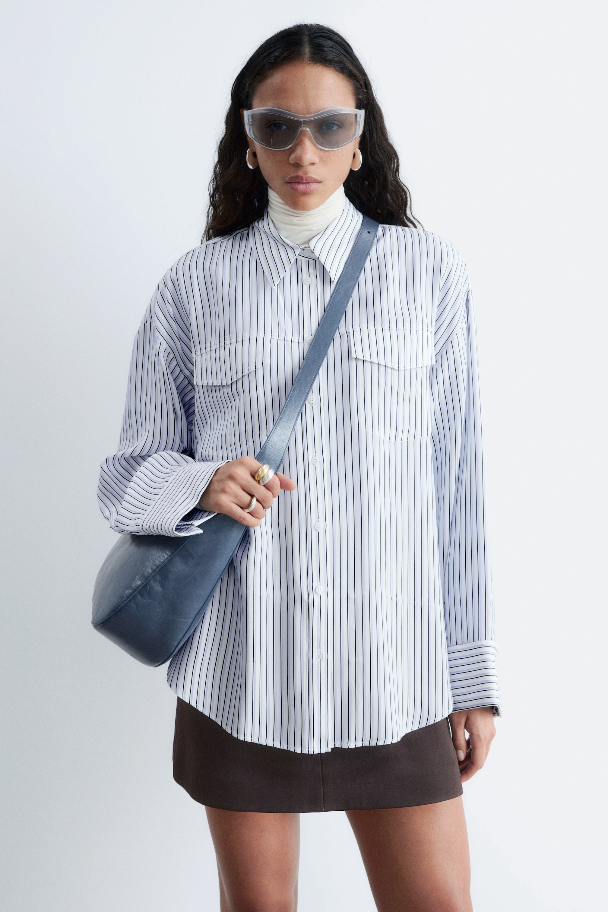 Ver imagen más grande: Una camisa blanca de botones con finas rayas verticales oscuras se muestra desde un ángulo frontal, con un cuello en punta, dos bolsillos en el pecho con solapa y mangas largas con puños abotonados. Se lleva superpuesta sobre un jersey de cuello alto blanco, con su corte holgado cayendo sobre una falda marrón y un bolso de hombro azul cruzando por delante.