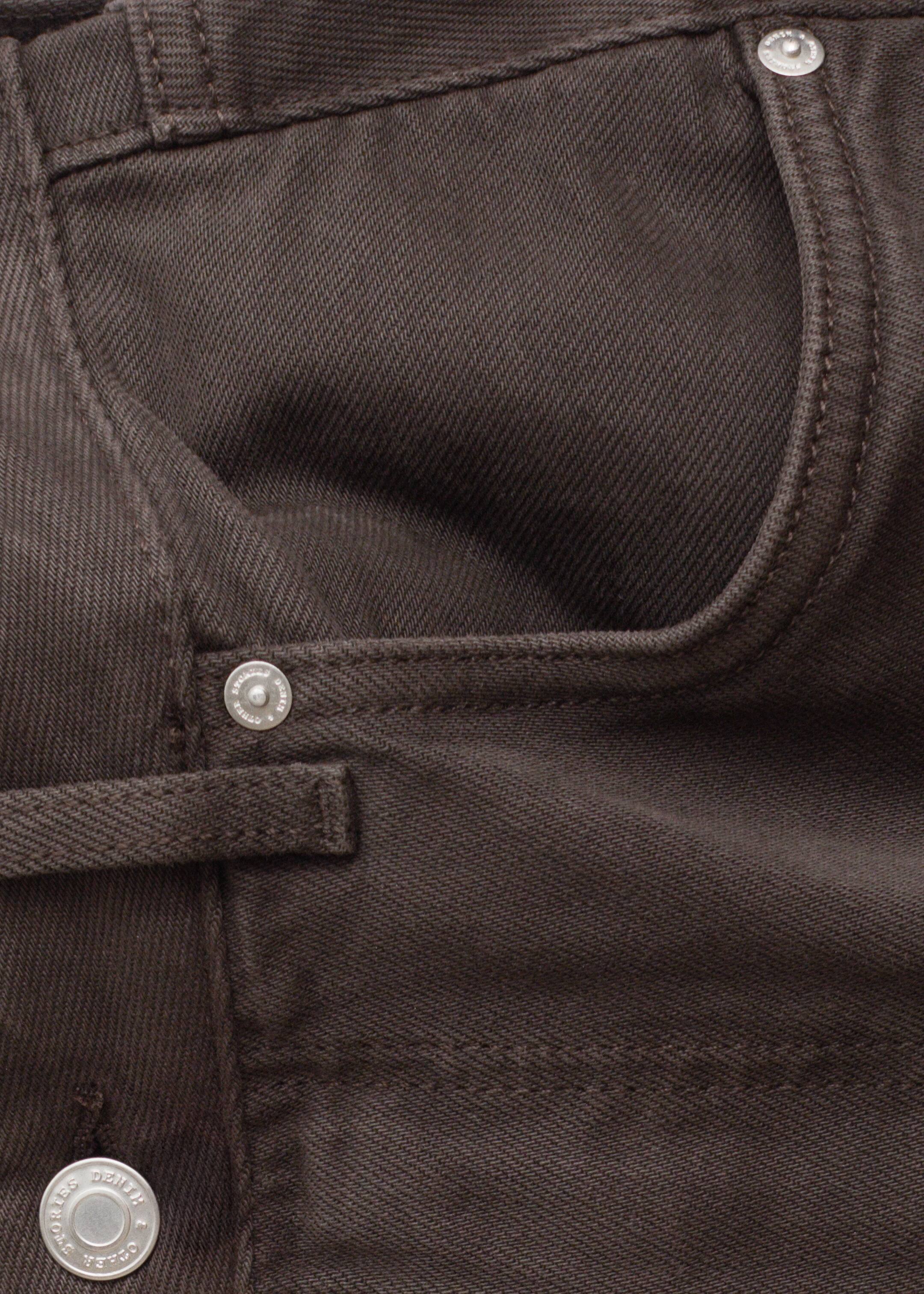 Grotere afbeelding bekijken: De voorkant van de bruine denim broek is zichtbaar, met een gebogen voorzak aan de rechterkant die is verstevigd met een zilverkleurige klinknagel. Een kleinere muntzak is zichtbaar binnenin de hoofdzak, ook verstevigd met een zilverkleurige klinknagel, en een riemlus loopt horizontaal aan de linkerkant.