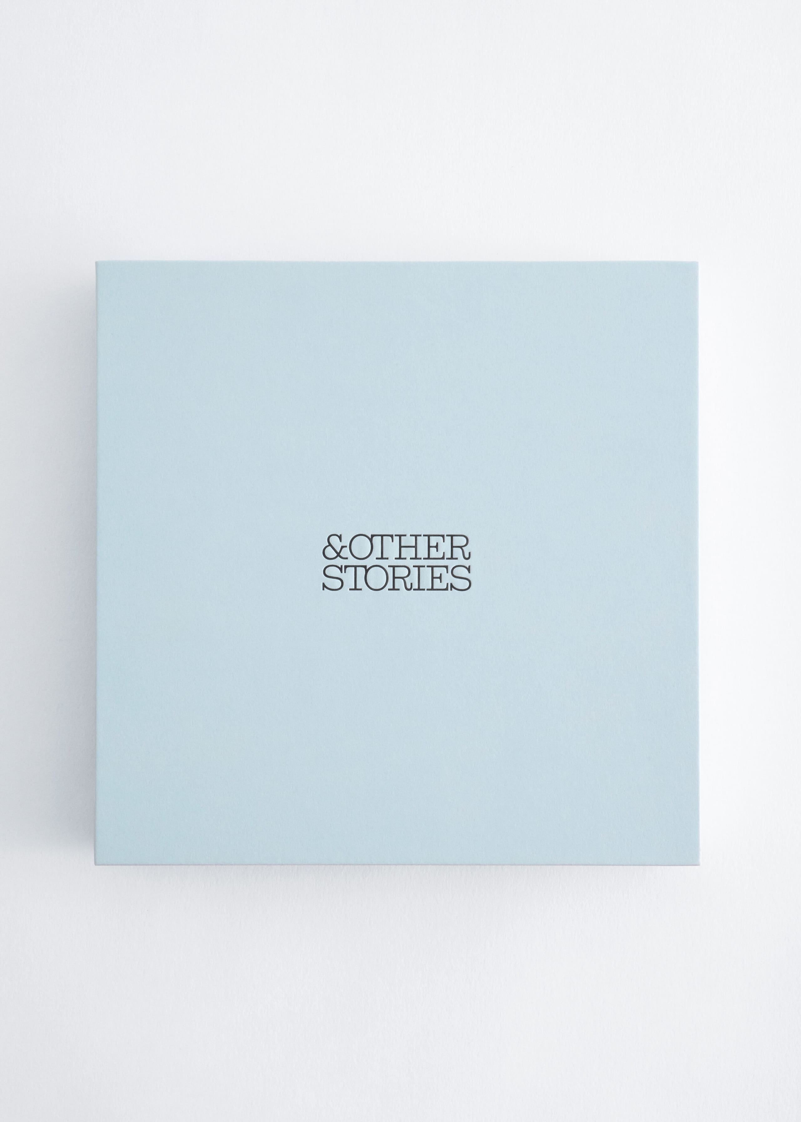 A square, light blue box is shown from the front, featuring the brand name "&OTHER STORIES" printed in a black serif font at the center of its smooth matte lid.