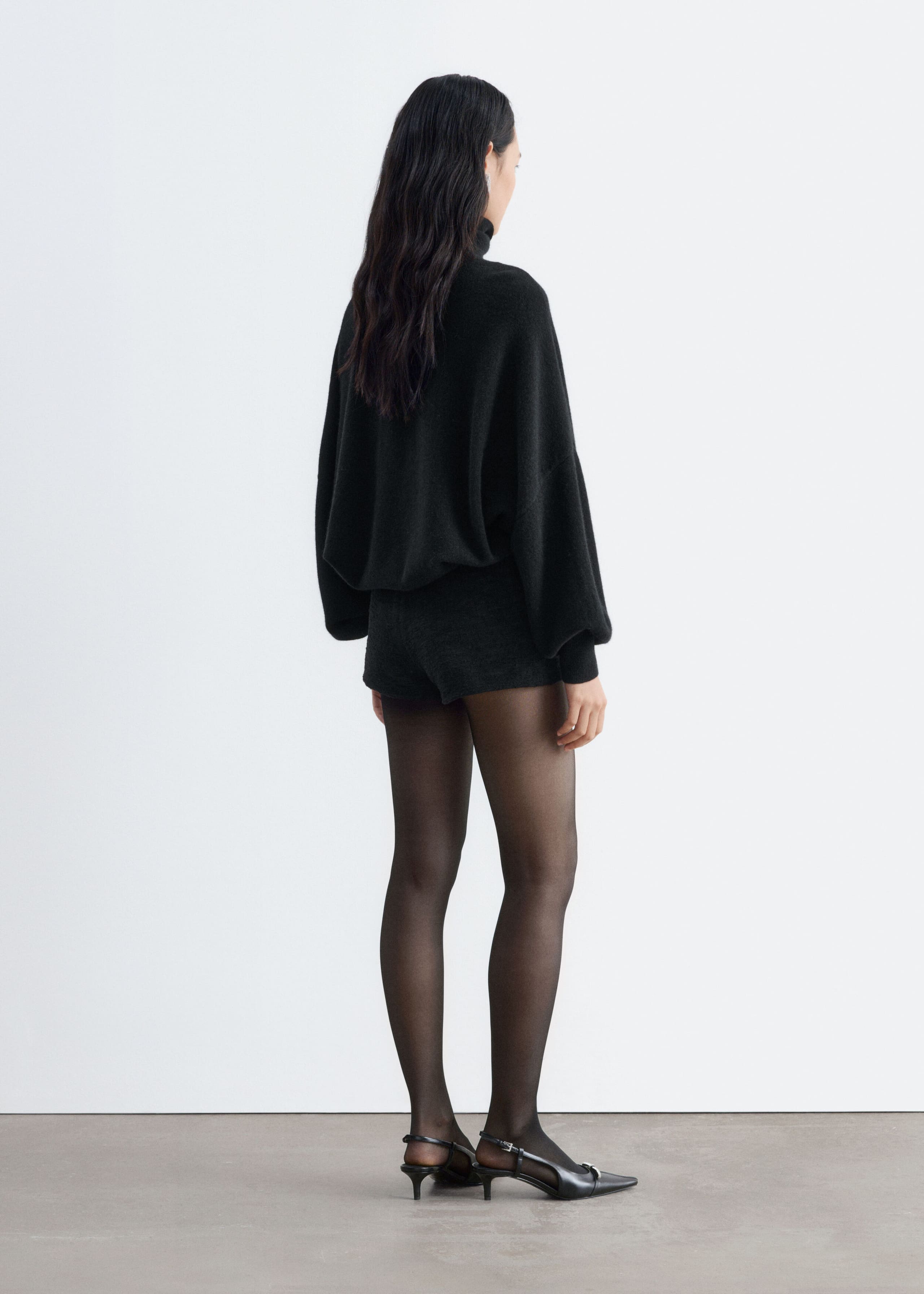 Black tweed shorts with a short, fitted silhouette are being worn by the model, shown from the rear. The shorts are styled with a loose black high-neck jumper, sheer black tights, and black pointed-toe slingback heels.