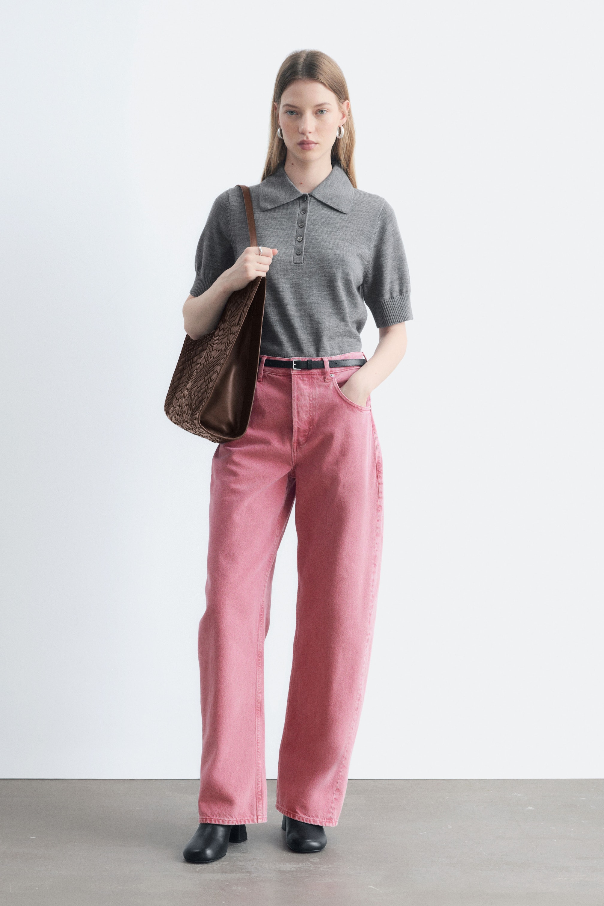 Agrandir l'image: Le mannequin porte un jean rose taille haute à jambe large, en position debout avec une main glissée dans la poche. Le jean est associé à un polo gris à manches courtes rentré, à une fine ceinture noire, à des bottes noires et à un sac à bandoulière tressé marron.