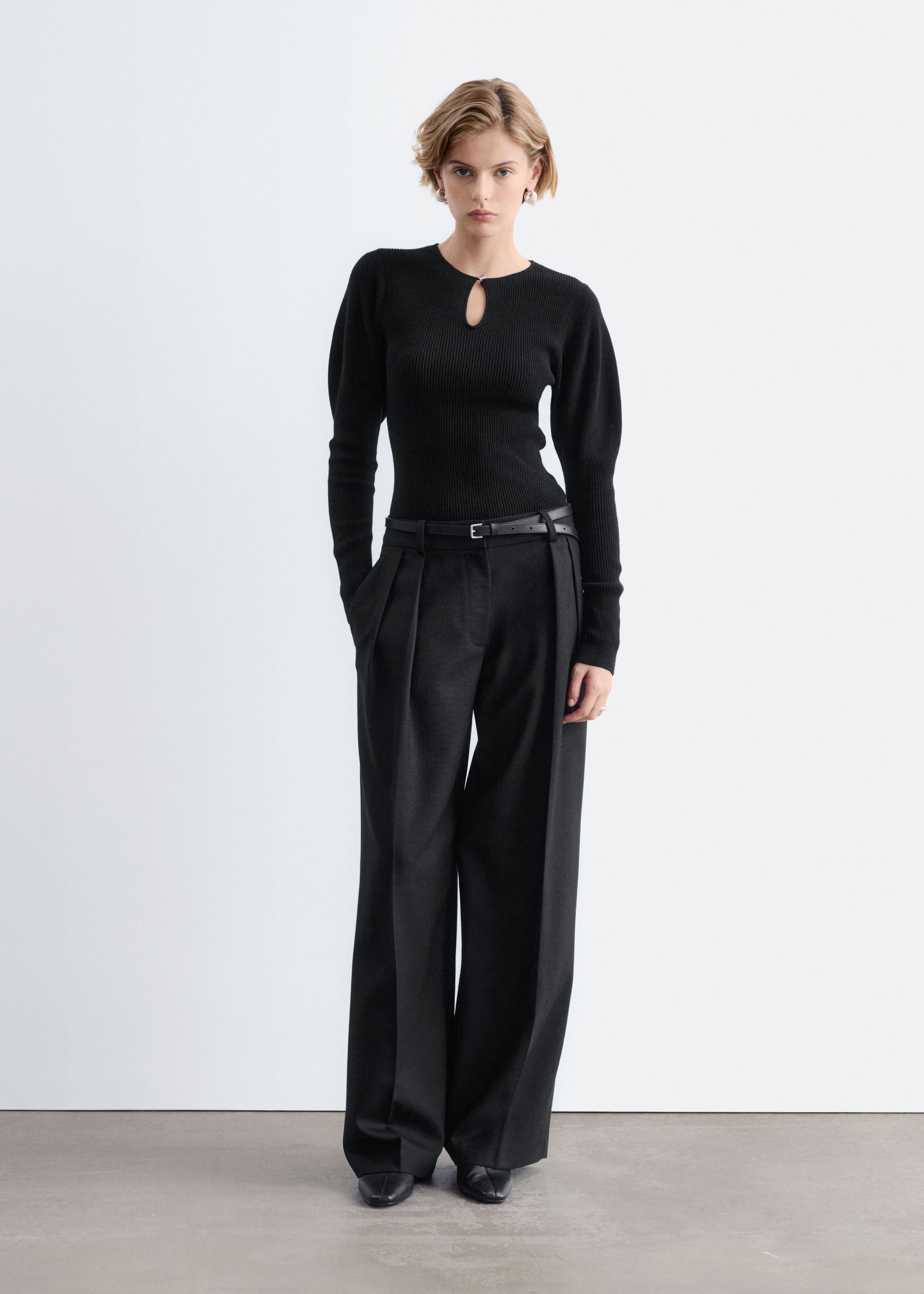 A black rib-knit long-sleeve top with a small keyhole cutout at the neckline and softly puffed shoulders is worn by the model. The slim-fit top is tucked into wide-leg black trousers styled with a thin black belt.