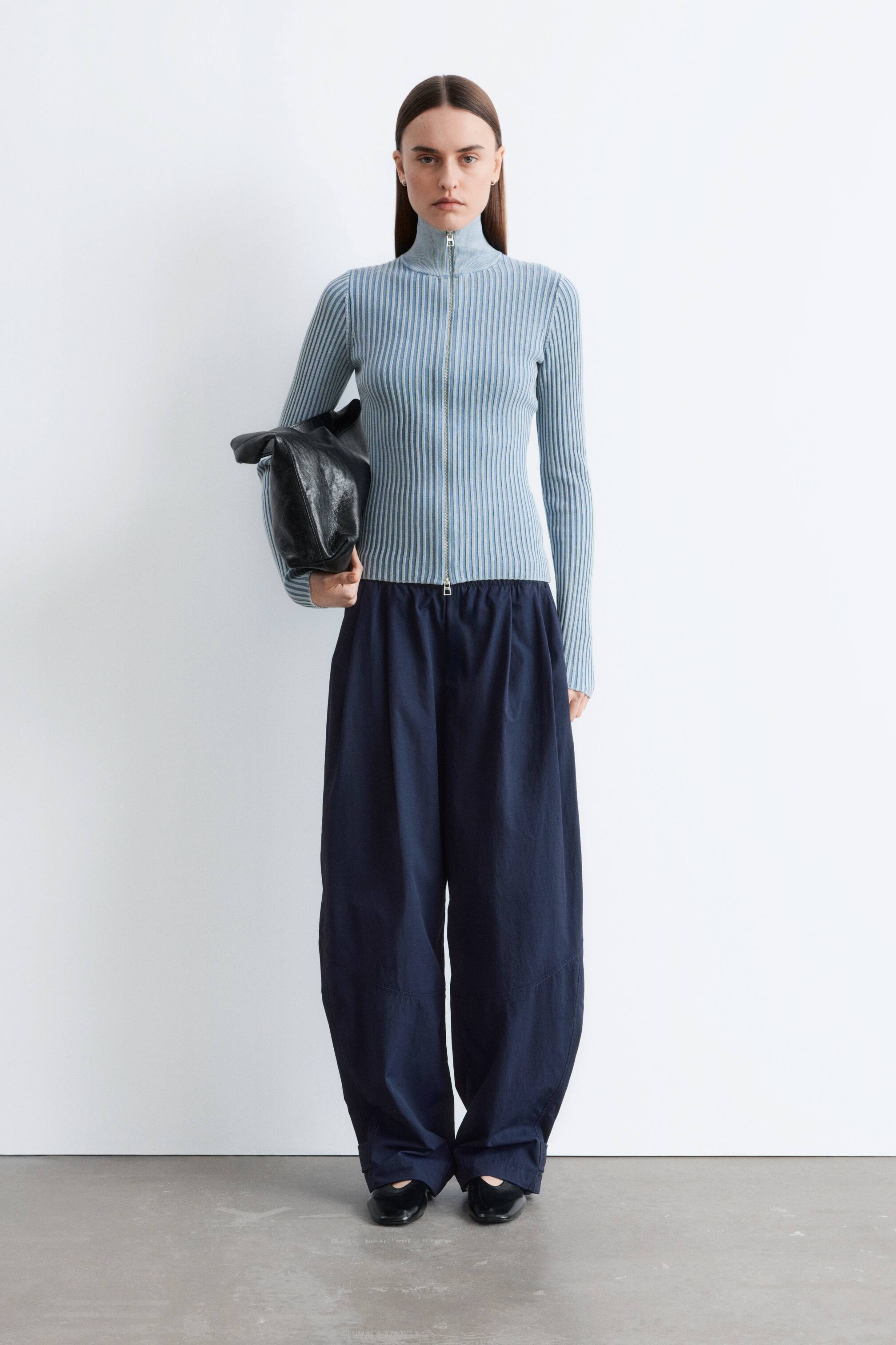 הצגת תמונה גדולה יותר: חולצת ריב סרוגה בצבע תכלת בהיר עם רוכסן מלא, צווארון גבוה ושרוולים ארוכים, מוצגת לצד מכנסיים רחבי-רגל בצבע נייבי כהה עם מותן בעל שרוך. תיק שחור רך ונפול מוחזק בצד, ונעליים שטוחות שחורות נראות מתחת למכנסיים.