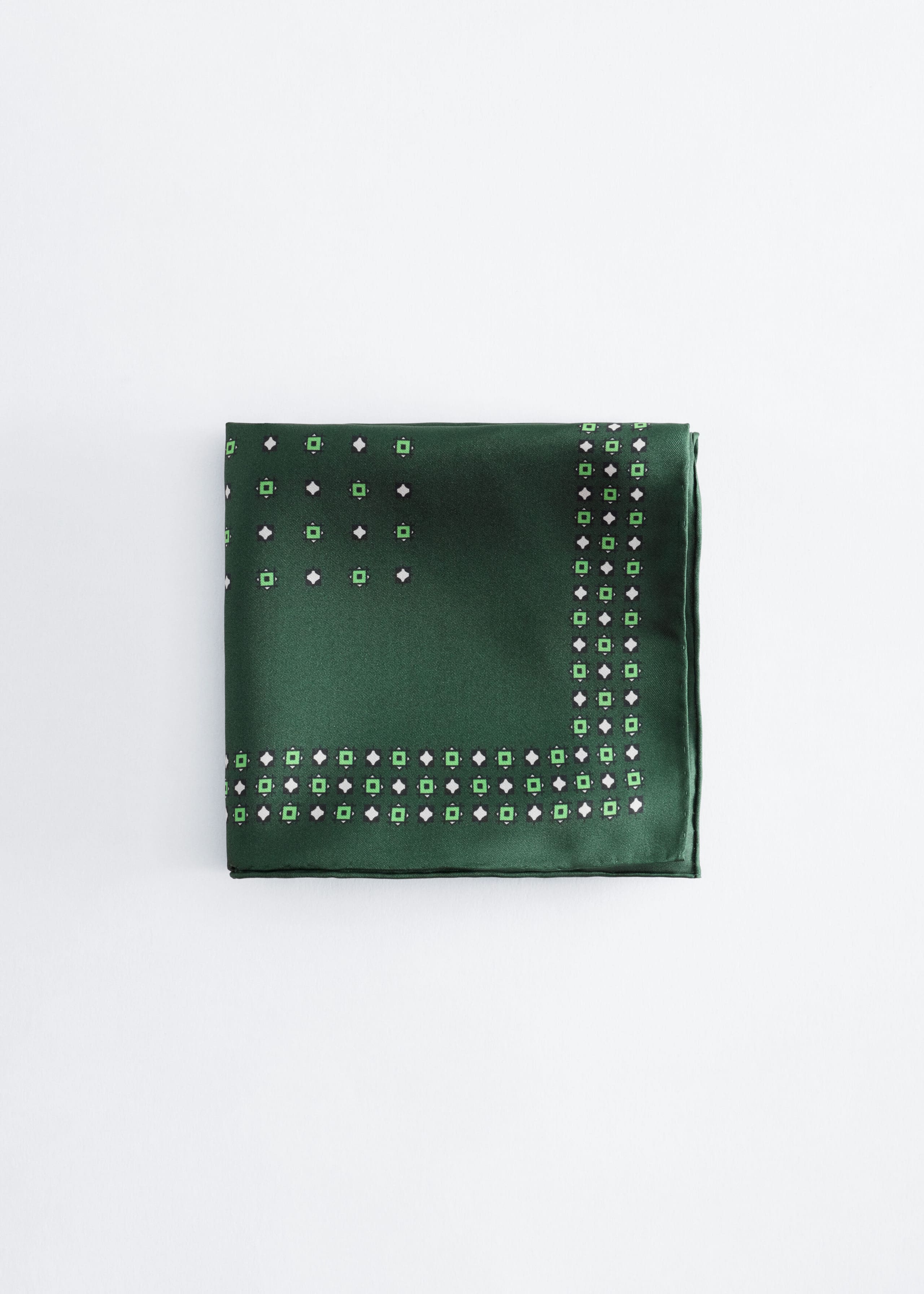 A green satin pocket square lies flat, facing forward, showcasing a geometric pattern of small white diamonds and green squares outlined in black. The pattern forms a dense border along the bottom and right edges and appears more sparsely in the top-left corner.