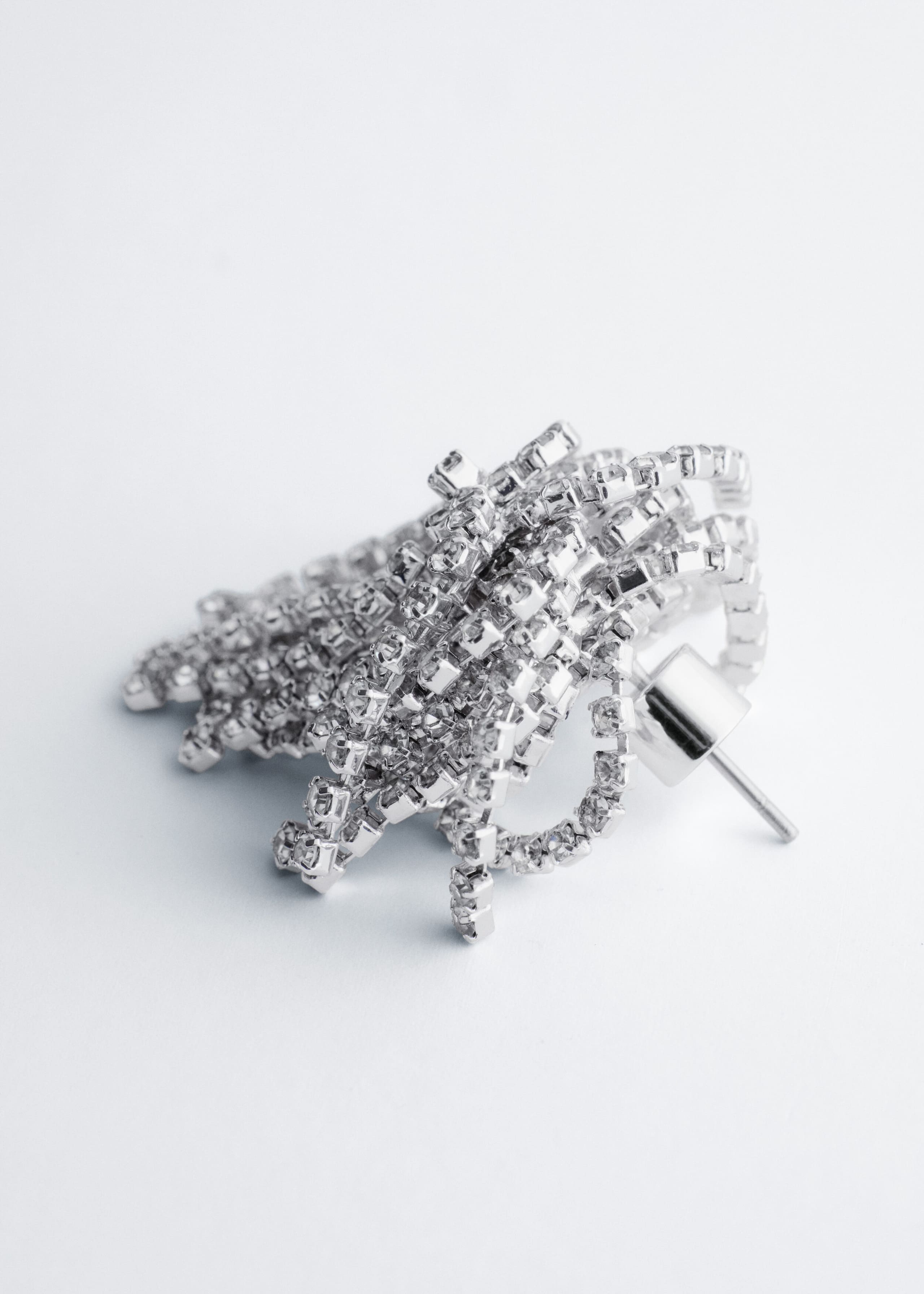A silver-toned brass earring featuring multiple looped strands of sparkling rhinestones is lying on its side, showing the post and cylindrical metal backing on the right. The dense cluster of rhinestone chains creates a voluminous, dimensional design.