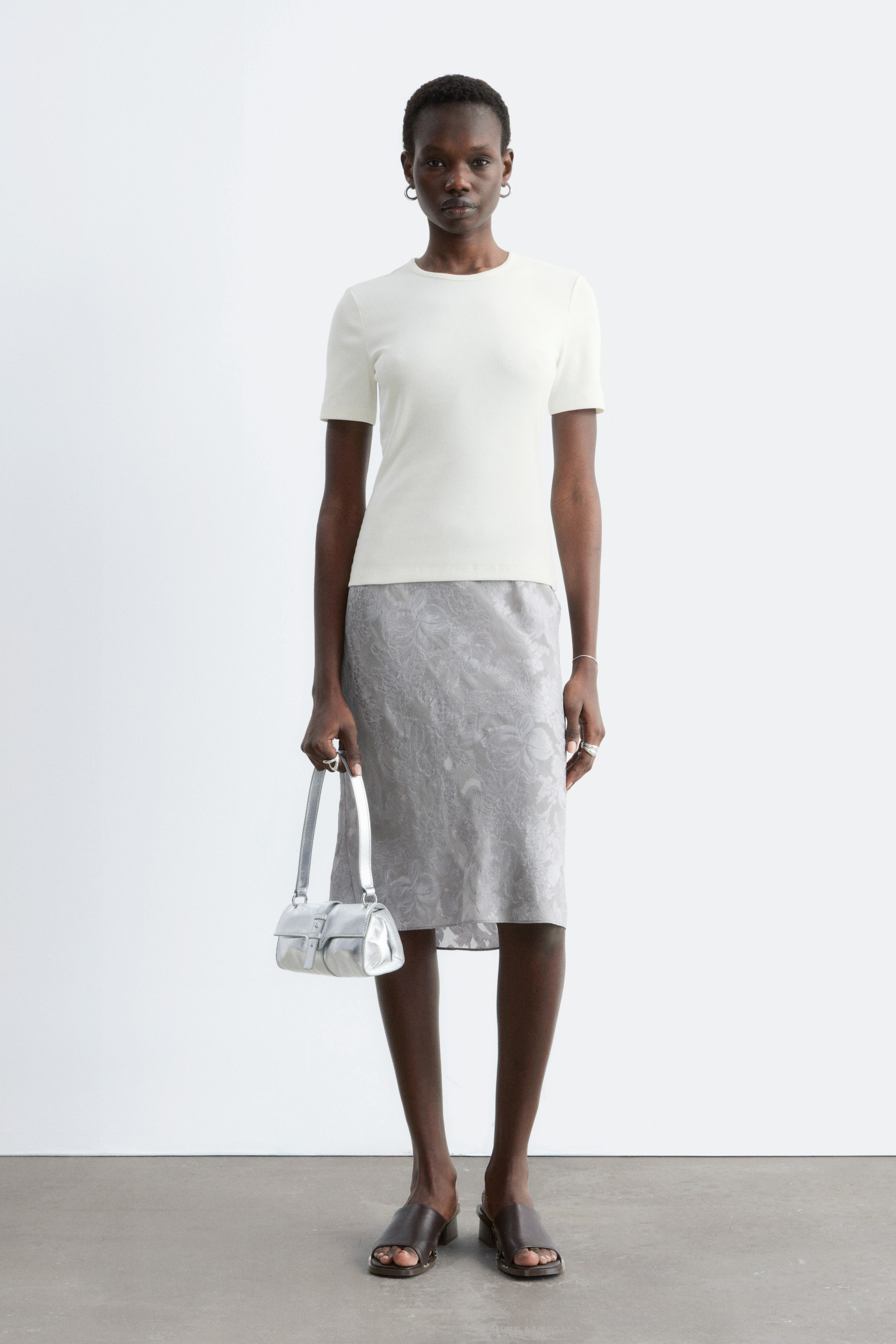 Visa större bild: En benvit, slim-fit T-shirt med rund hals och korta ärmar bärs av modellen, stylad med en gråmönstrad midikjol.