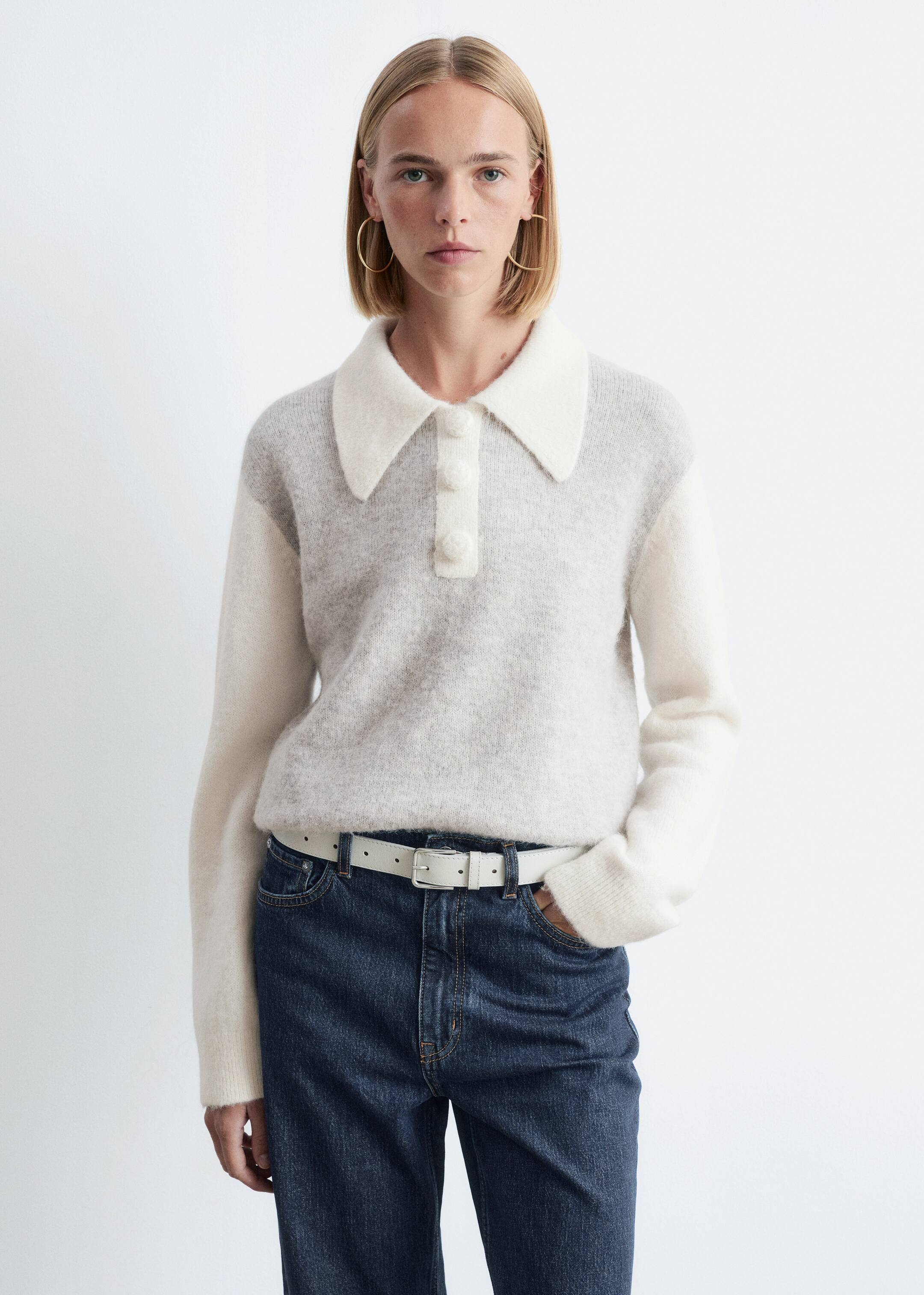 Ver imagen más grande: Un jersey de punto bicolor, con un cuerpo gris claro, mangas largas blancas con puños acanalados y un cuello blanco oversize con una tapeta decorativa y tres botones cubiertos, está siendo usado por una modelo. La modelo está de pie, con una mano en el bolsillo de unos vaqueros oscuros, que están combinados con un cinturón blanco.