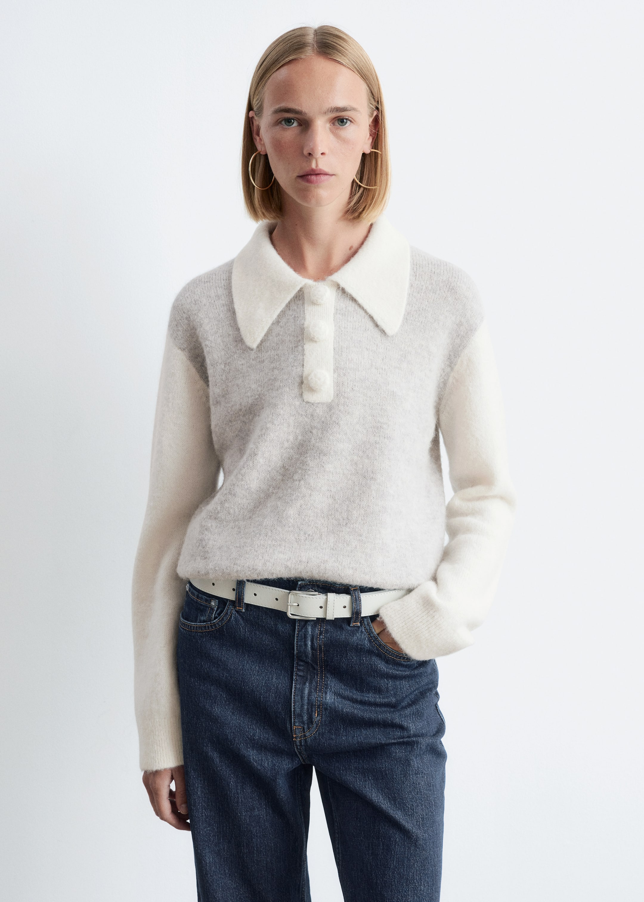 Grösseres Bild ansehen: Ein zweifarbiger Strickpullover, der einen hellgrauen Körper, weiße lange Ärmel mit gerippten Bündchen und einen übergroßen weißen Kragen mit einer dekorativen Knopfleiste und drei überzogenen Knöpfen aufweist, wird von einem Model getragen. Das Model steht mit einer Hand in der Tasche einer dunklen Jeans, die mit einem weißen Gürtel gestylt ist.
