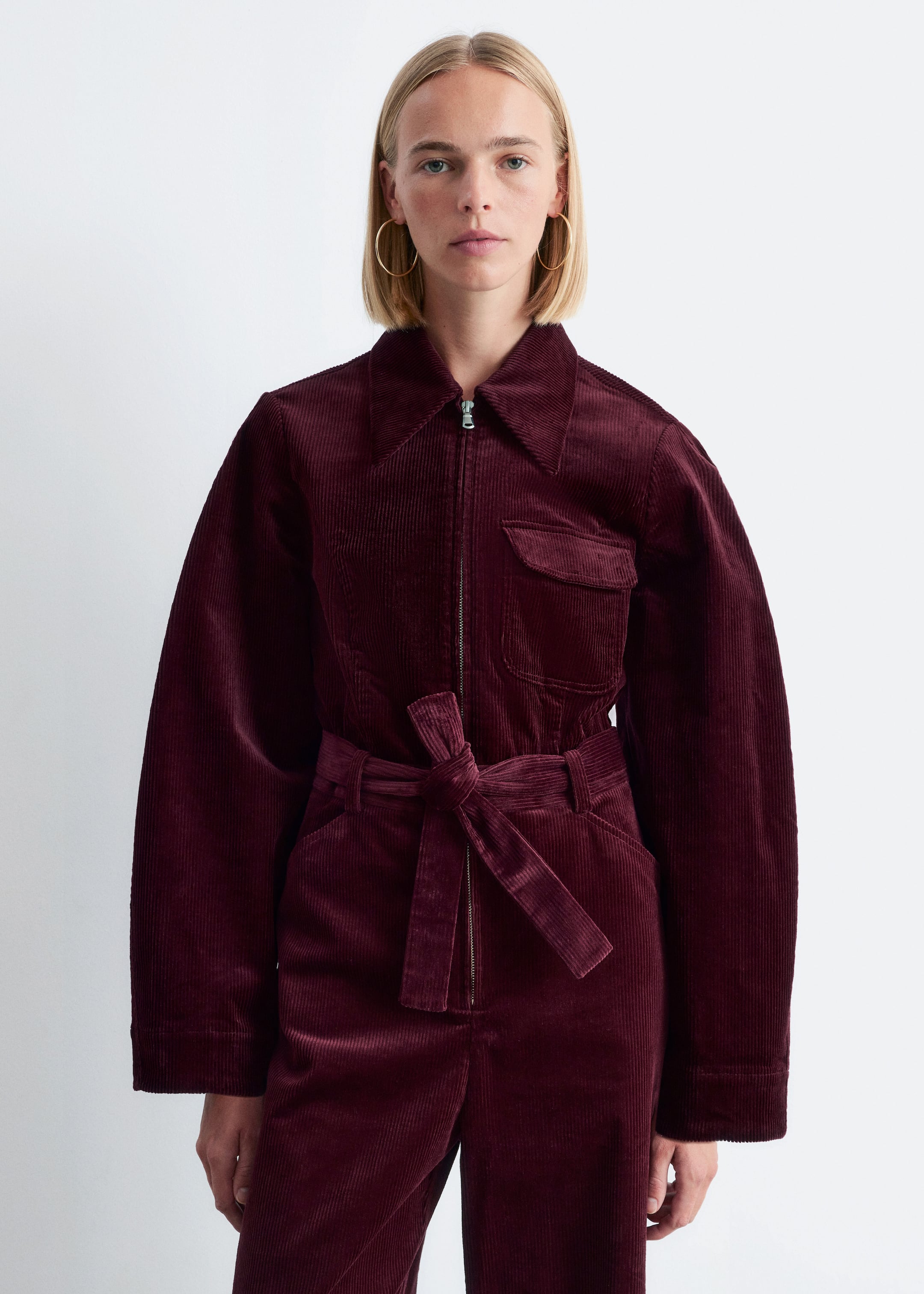 Größeres Bild anzeigen: Ein burgunderfarbener Cord-Jumpsuit wird aus einem frontalen Winkel präsentiert, mit einem durchgehenden Reißverschluss vorne, einem spitzen Kragen und voluminösen langen Ärmeln. Er ist mit einer einzelnen aufgesetzten Brusttasche, Seitentaschen im unteren Bereich und einem Gürtel aus demselben Stoff, der in der Taille gebunden wird, gestaltet.