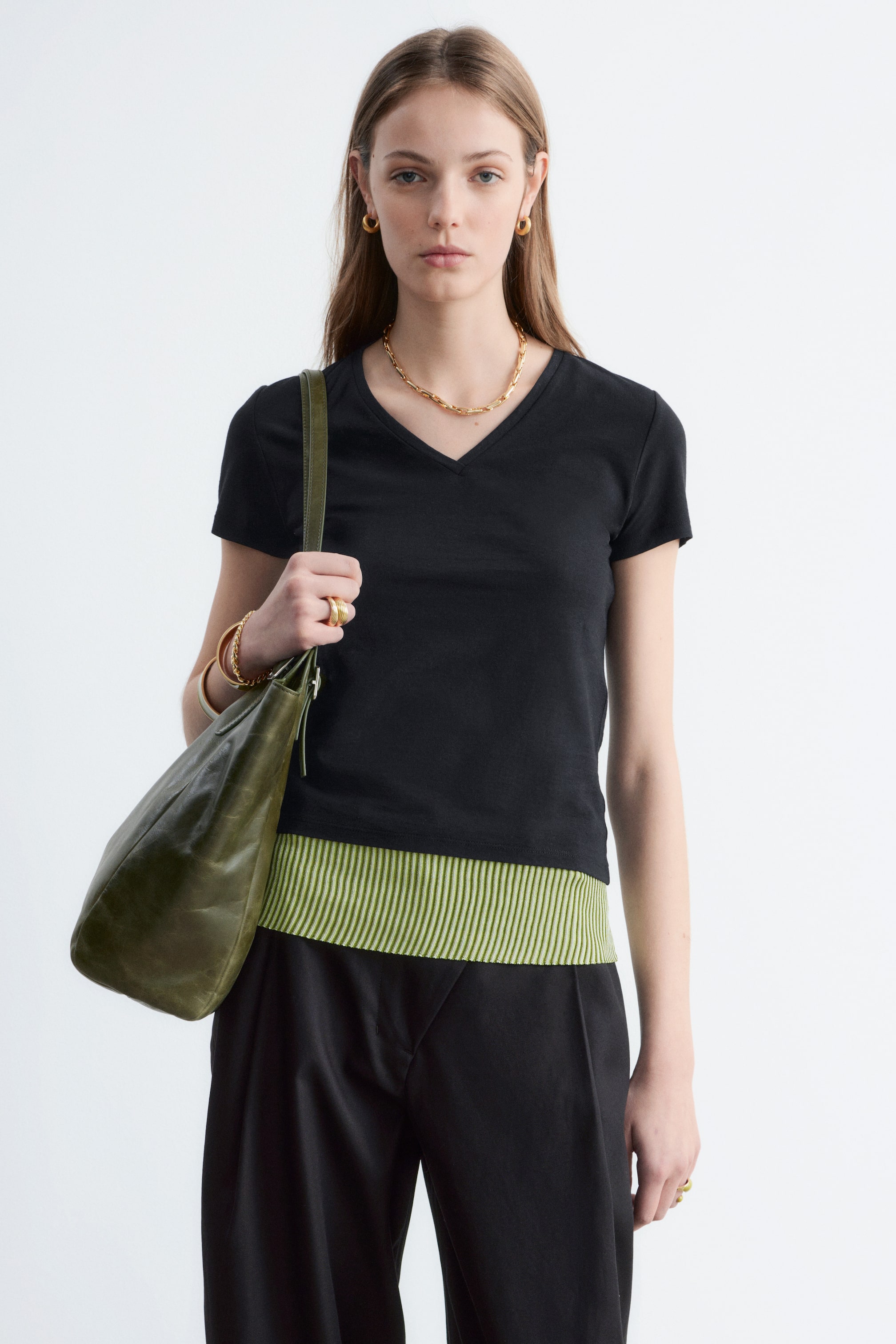 Zobrazit větší obrázek: Modelka má na sobě černé tričko s výstřihem do V a krátkými rukávy. Tričko je přiléhavé a stylově vrstvené přes zeleno-bílé pruhované top, doplněné černými kalhotami.