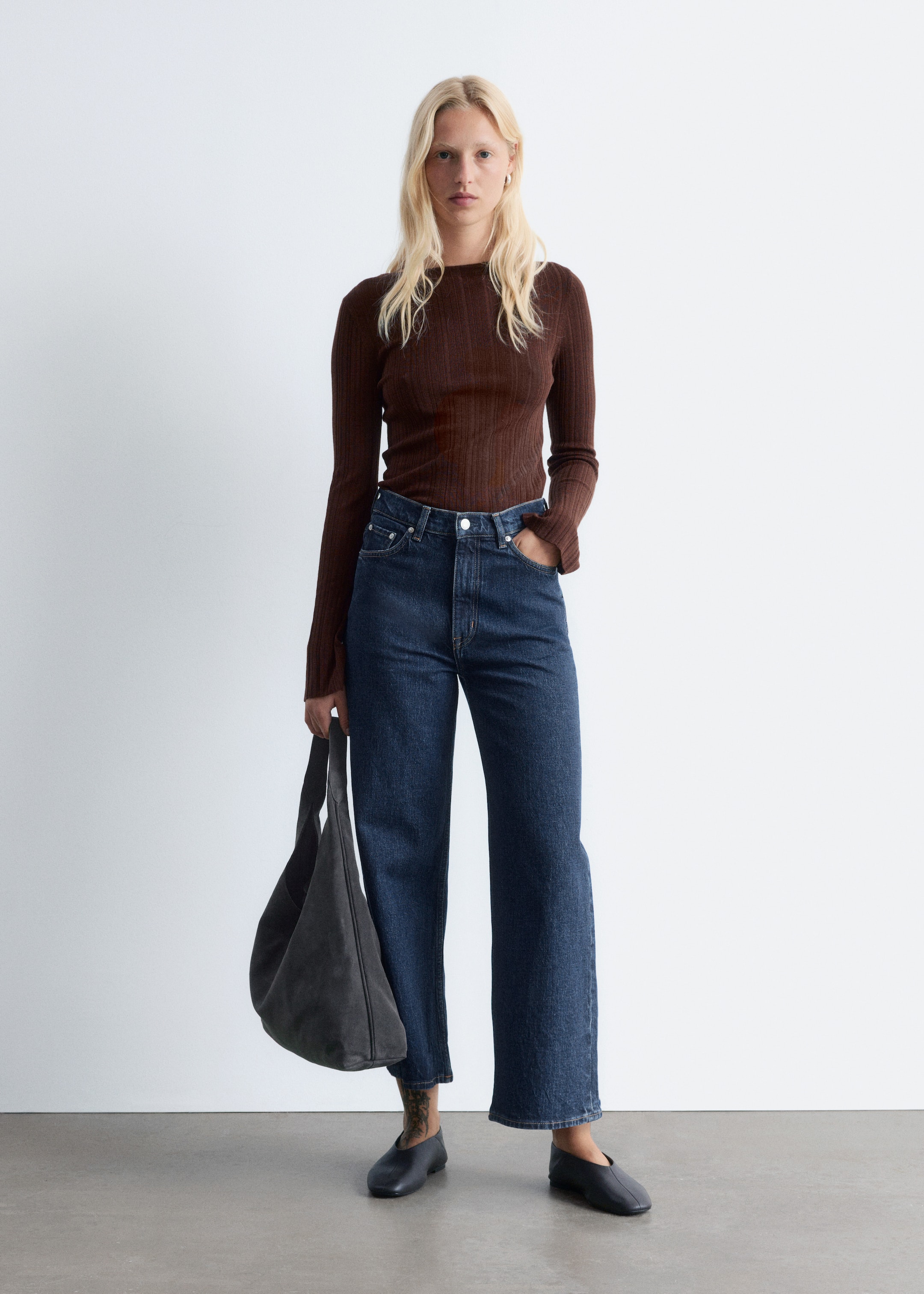 Visualizza immagine più grande: Un top a maniche lunghe in maglia a coste marrone scuro con scollo rotondo è indossato, abbinato a jeans in denim blu scuro a vita alta, a gamba larga e leggermente corti. Una borsa a spalla morbida e slouchy grigio scuro è portata, e delle scarpe piatte e scivolose scure completano l'insieme.