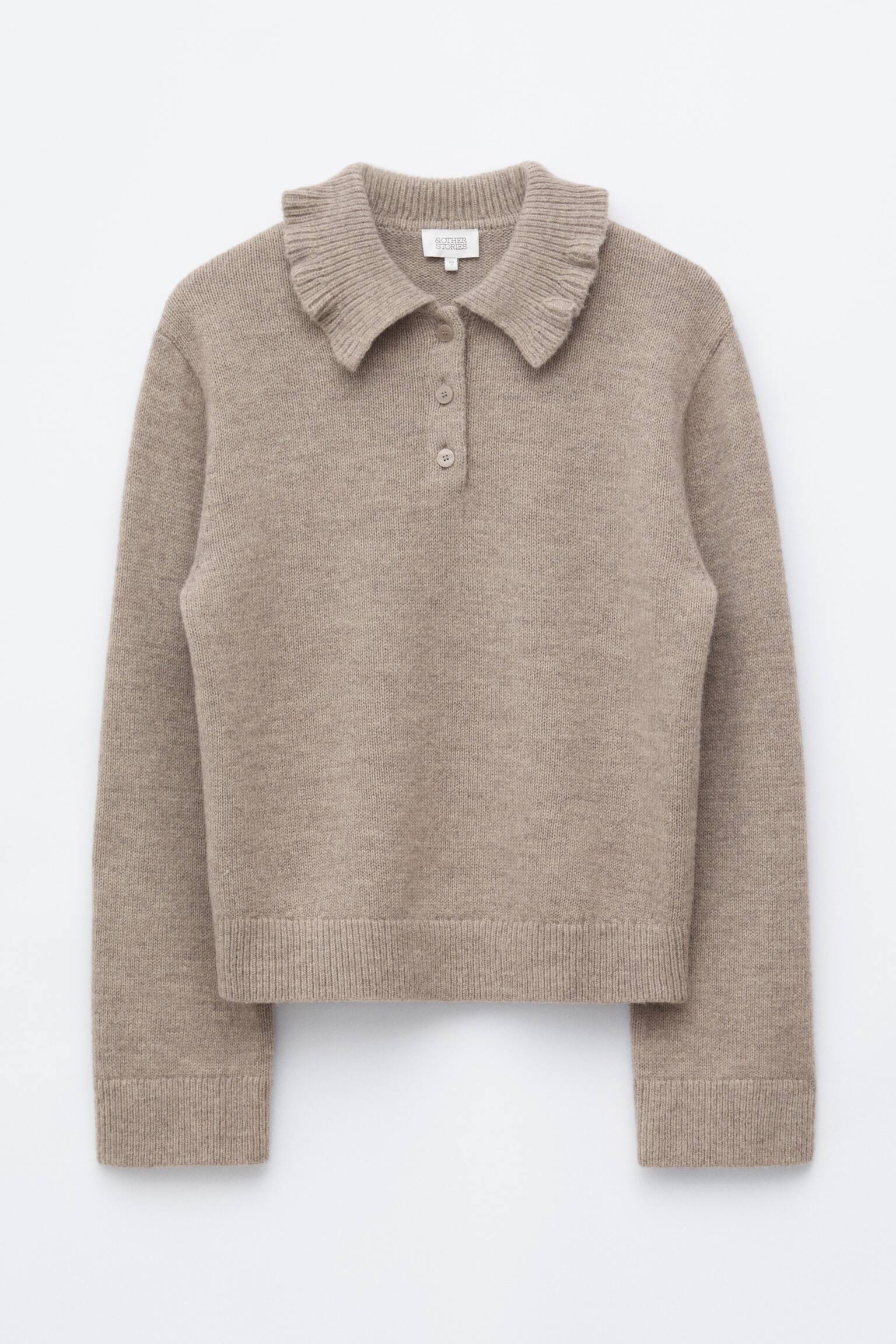 En beige jumper med volangkrage Ã¤r vÃ¤nd framÃ¥t och visar upp en bred, ribbad volangkrage och en knappslÃ¥ med tre knappar. Den lÃ¥ngÃ¤rmade stickade jumpren har ribbade muddar och en matchande ribbad nederkant.
