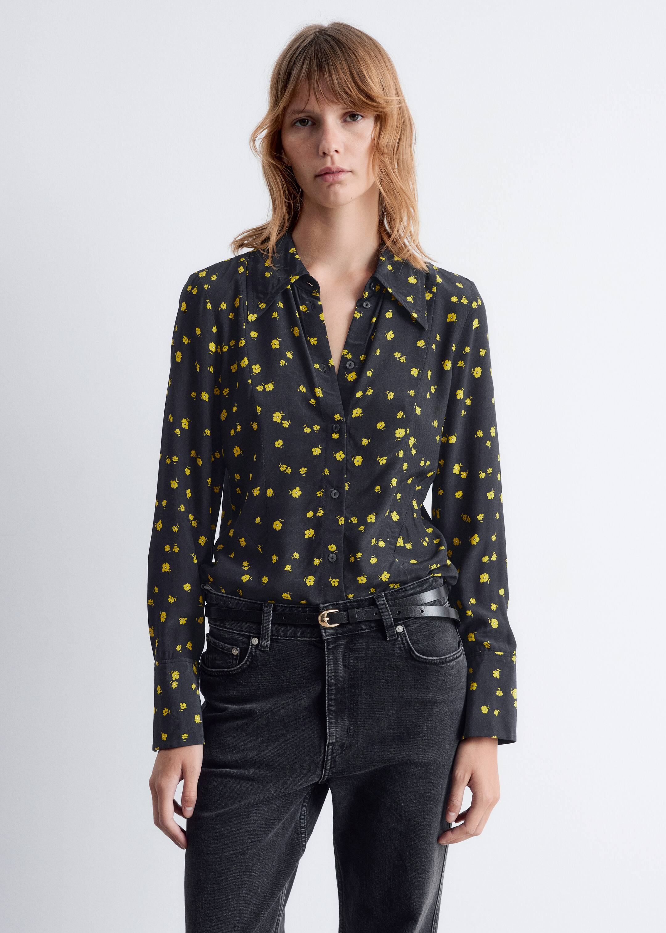 Ver imagem maior: Uma camisa preta de botões com um padrão floral amarelo está a ser usada. A camisa de mangas compridas apresenta uma gola pontiaguda e está enfiada num par de jeans pretos de cintura alta, com um cinto preto e uma fivela dourada visível na cintura.
