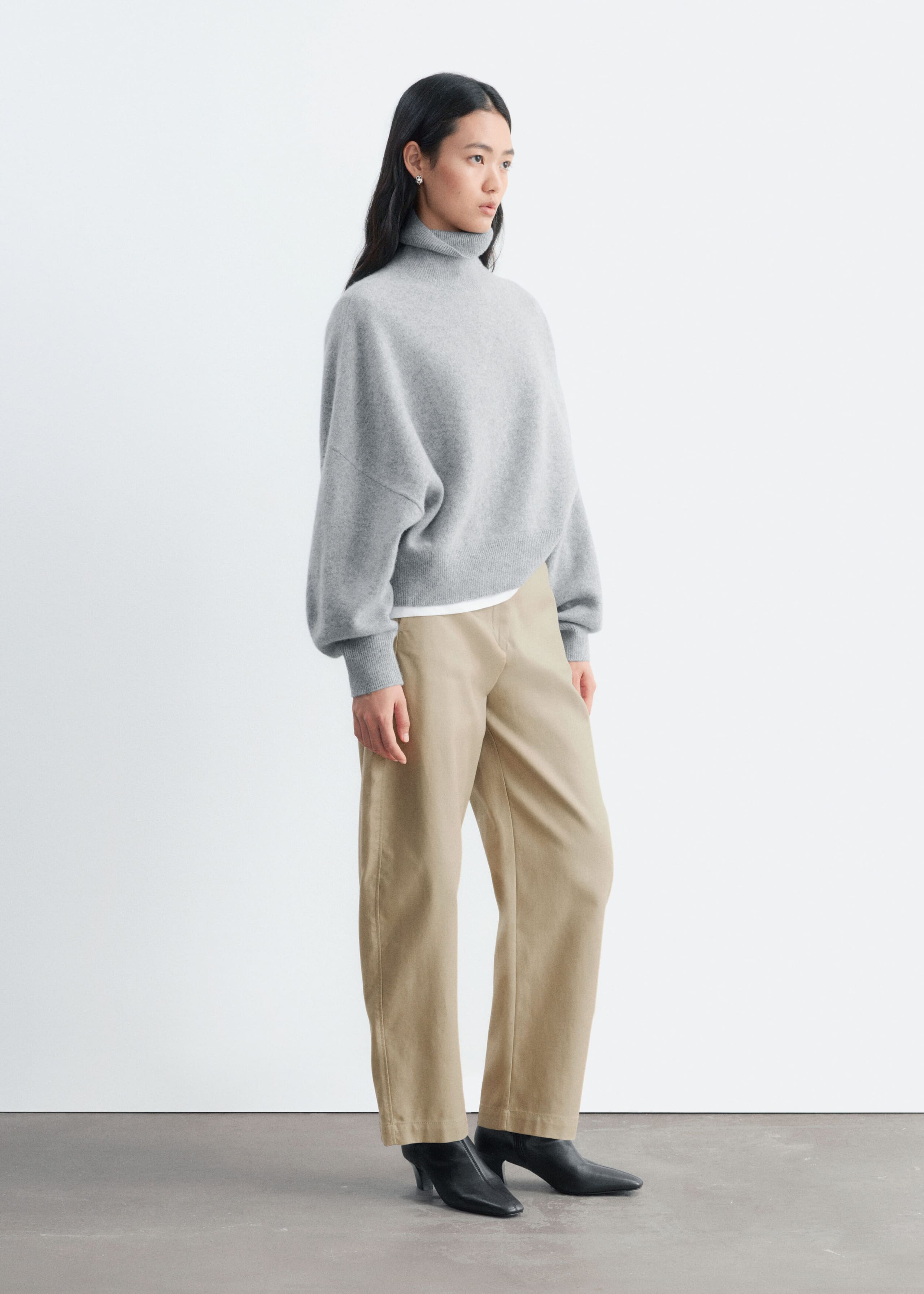 An oversized grey knit jumper featuring a folded rib turtleneck and lowered armholes is being worn by the model. The jumper is styled with beige trousers and a white undershirt visible at the hem.