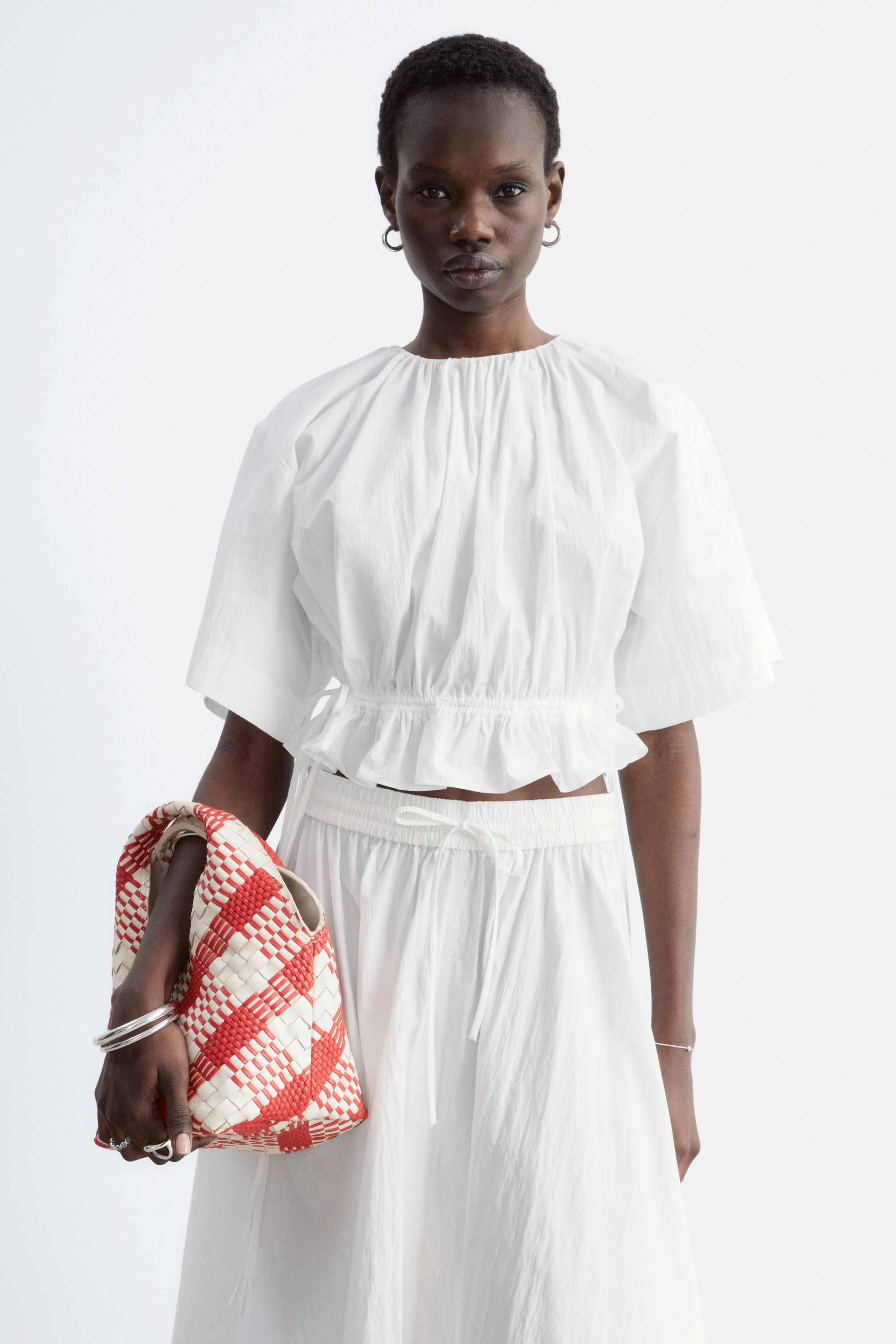 Visualizza l'immagine ingrandita: Il top bianco corto presenta uno scollo rotondo arricciato e maniche corte e voluminose. È dotato di un orlo elasticizzato e arricciato che si trova in vita, e il top è rivolto in avanti, mostrando la sua vestibilità rilassata.