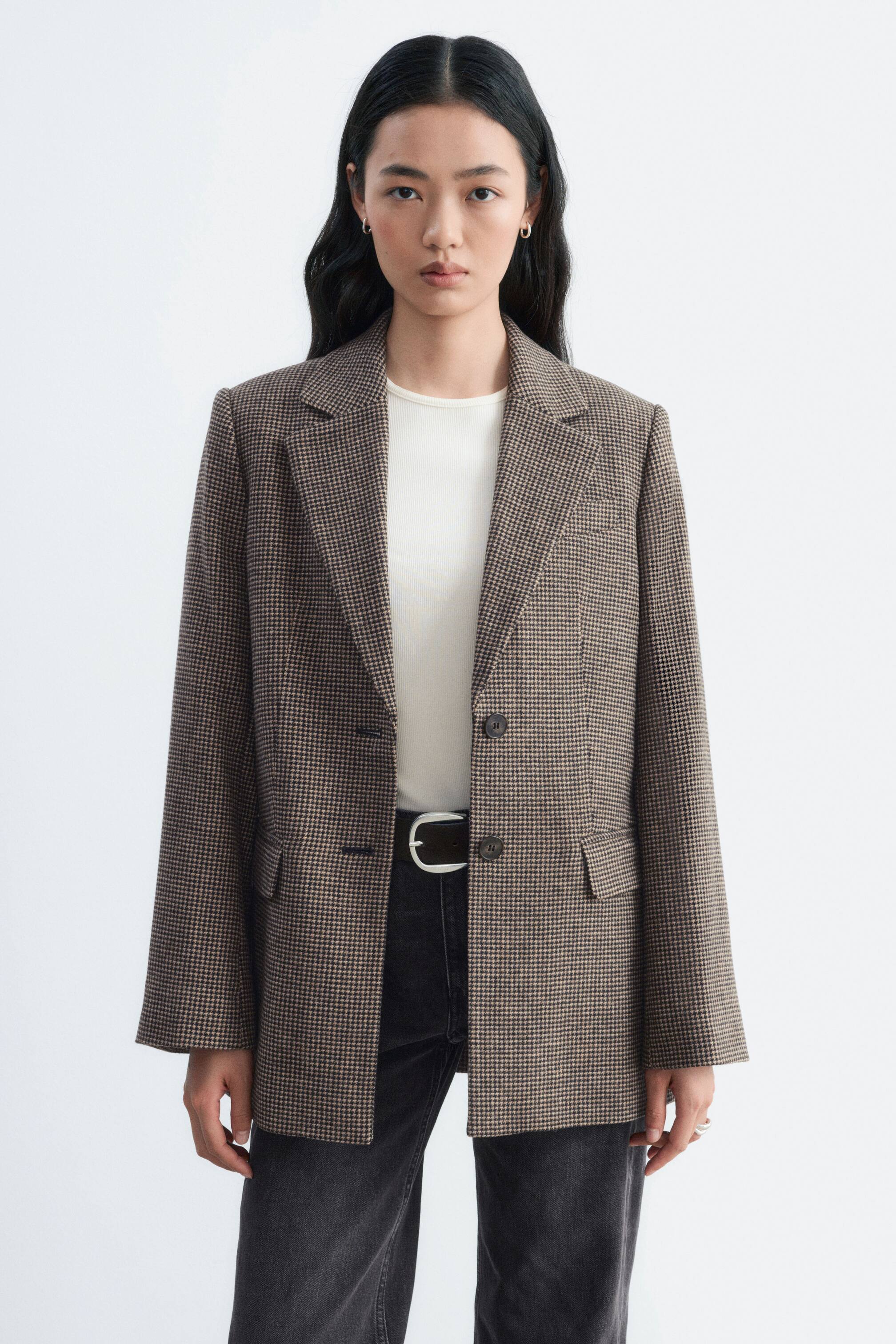 Agrandir l'image: Un blazer à pied-de-poule marron est porté ouvert par le modèle, superposé sur un haut blanc à col rond et un jean gris foncé. Ce blazer à simple boutonnage présente des épaules structurées, des revers crantés et des poches à rabat sur le devant.