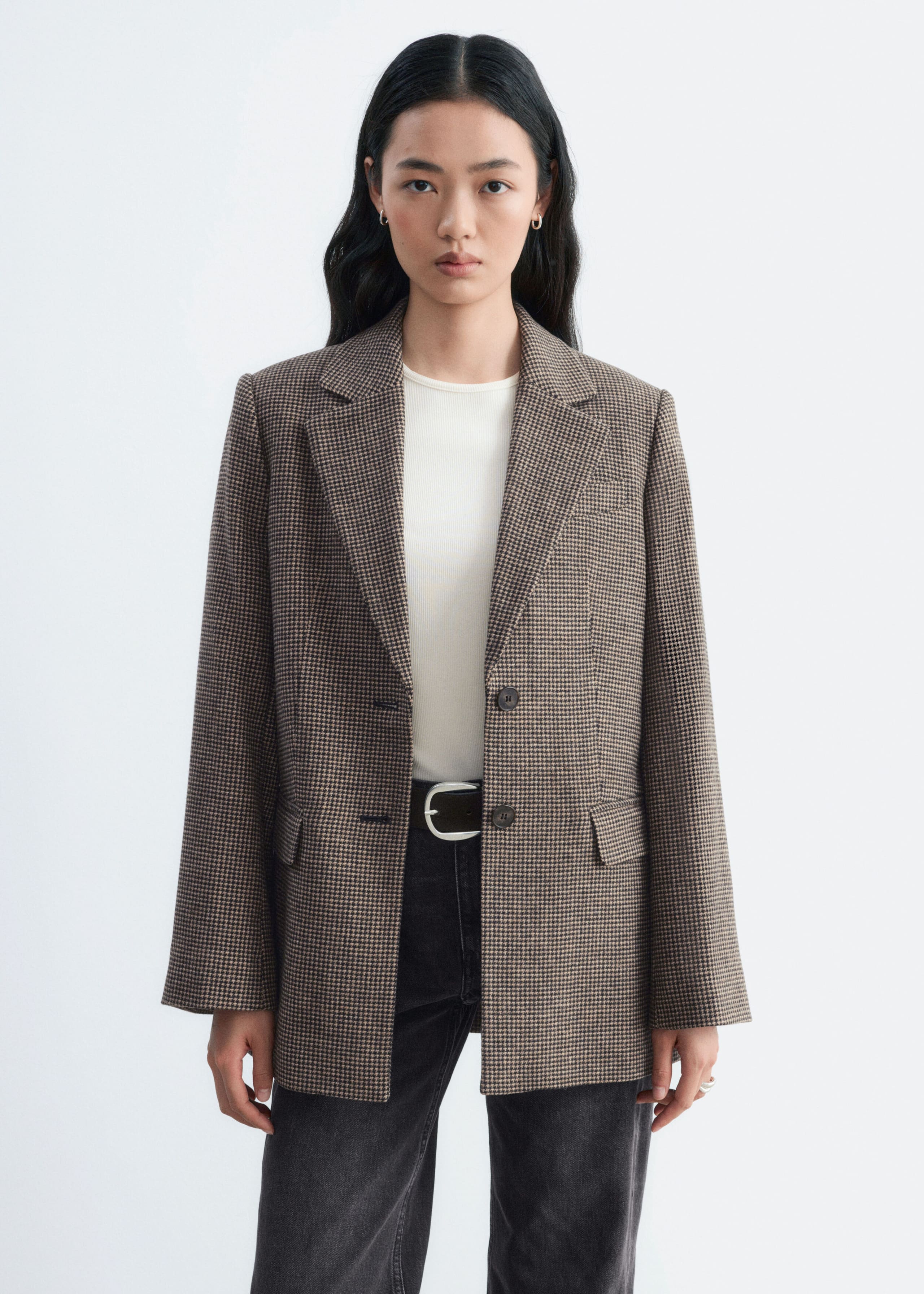 A brown houndstooth blazer is being worn open by the model, layered over a white crew neck top and dark grey jeans. The single-breasted blazer features structured shoulders, notch lapels, and front flap pockets.