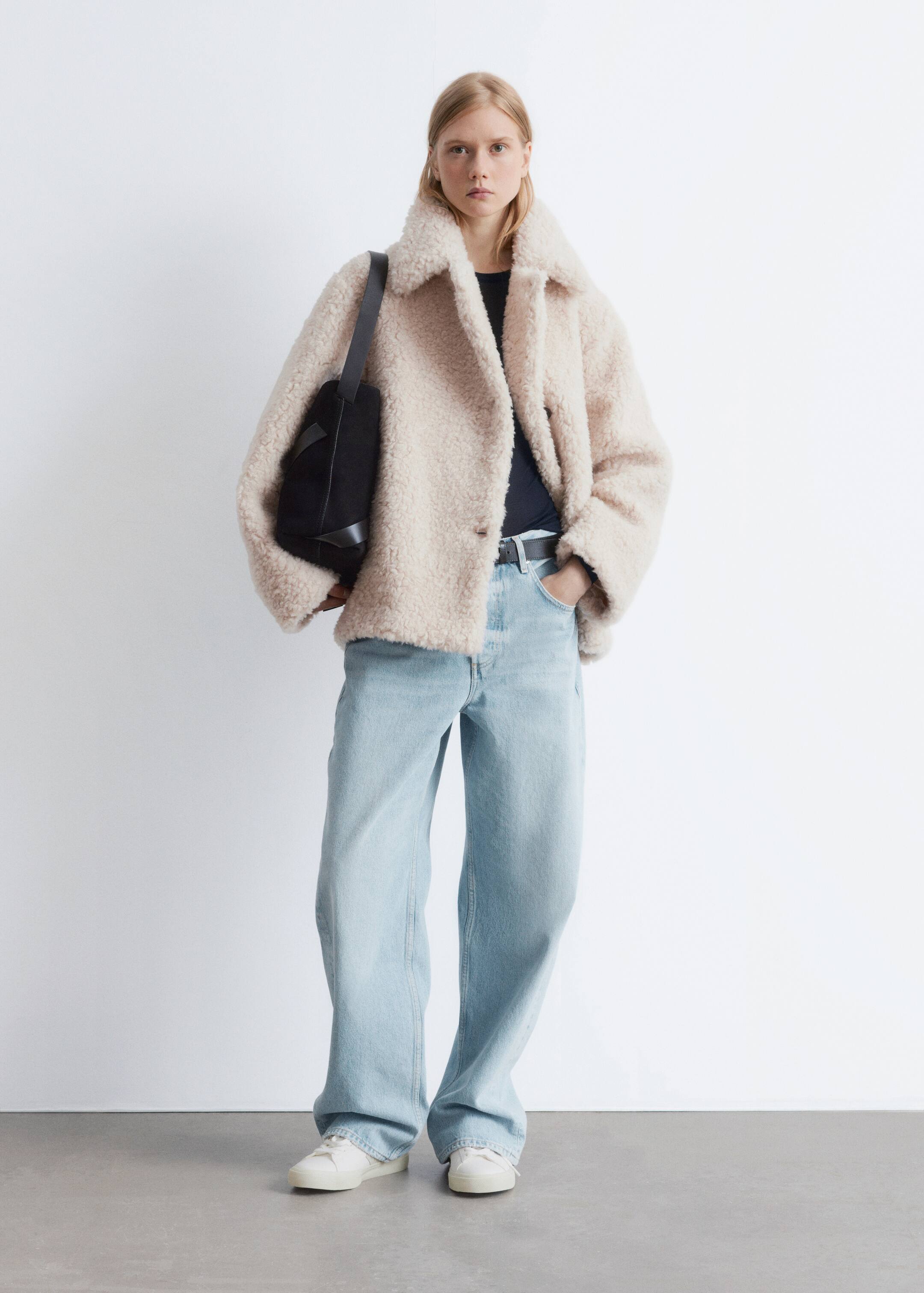 Προβολή μεγαλύτερης εικόνας: Ένα κρεμ μπουφάν tedd y φοριέται ανοιχτό πάνω από ένα μαύρο μπλουζάκι, συνδυασμένο με τζιν φαρδιάς γραμμής και λευκά αθλητικά παπούτσια, παρουσιασμένο από μπροστινή γωνία. Μια μαύρη ζώνη σφίγγει τη μέση και μία μαύρη τσάντα ώμου κρέμεται από τον αριστερό ώμο.