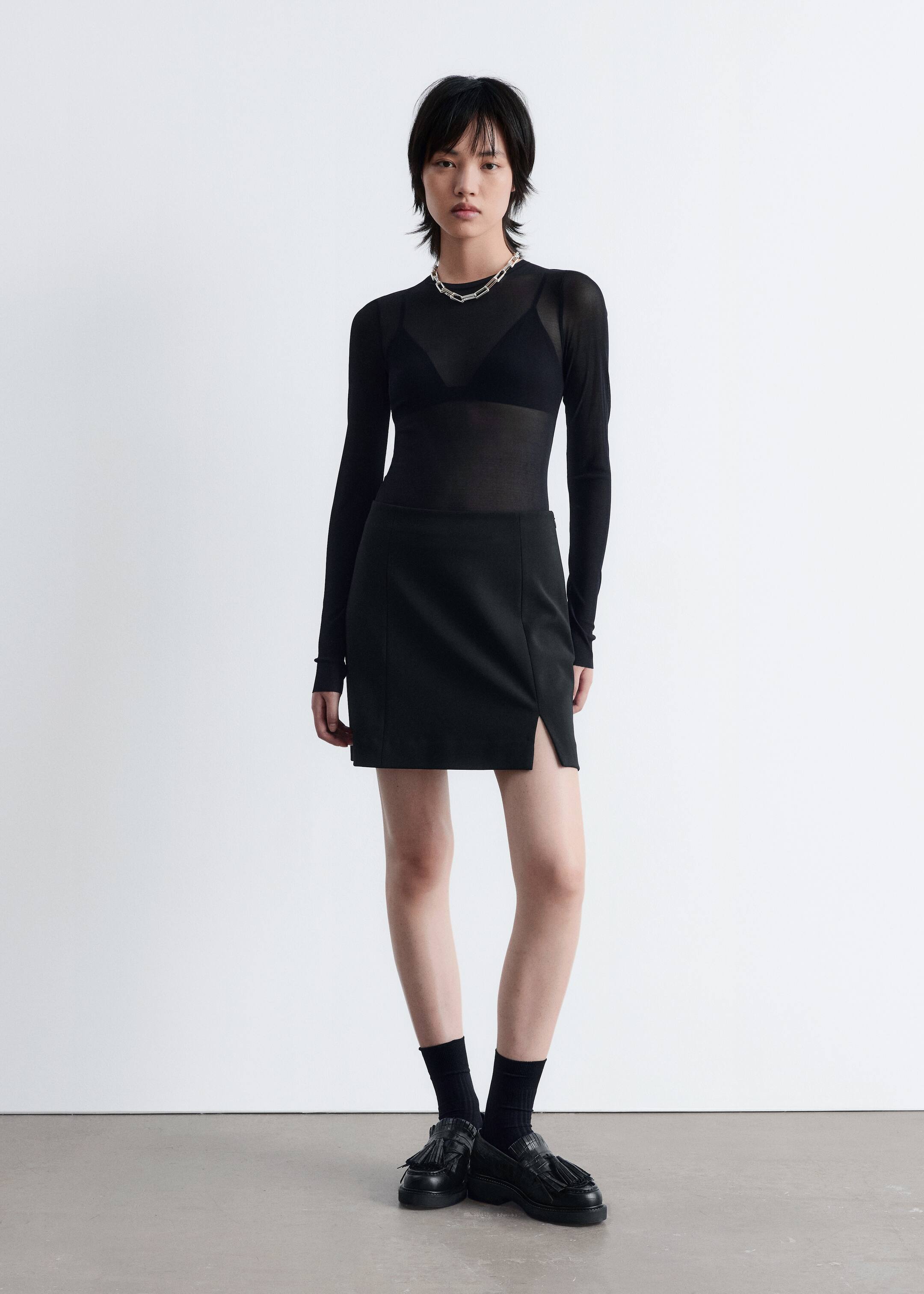 Agrandir l'image: Un haut noir transparent à manches longues est porté par-dessus un bralette noir, accessoirisé d'un collier à maillons en argent. Une mini-jupe noire avec une fente sur le côté est portée. Des mocassins noirs à glands avec une finition texturée complètent l'ensemble, portés avec des chaussettes noires montantes.